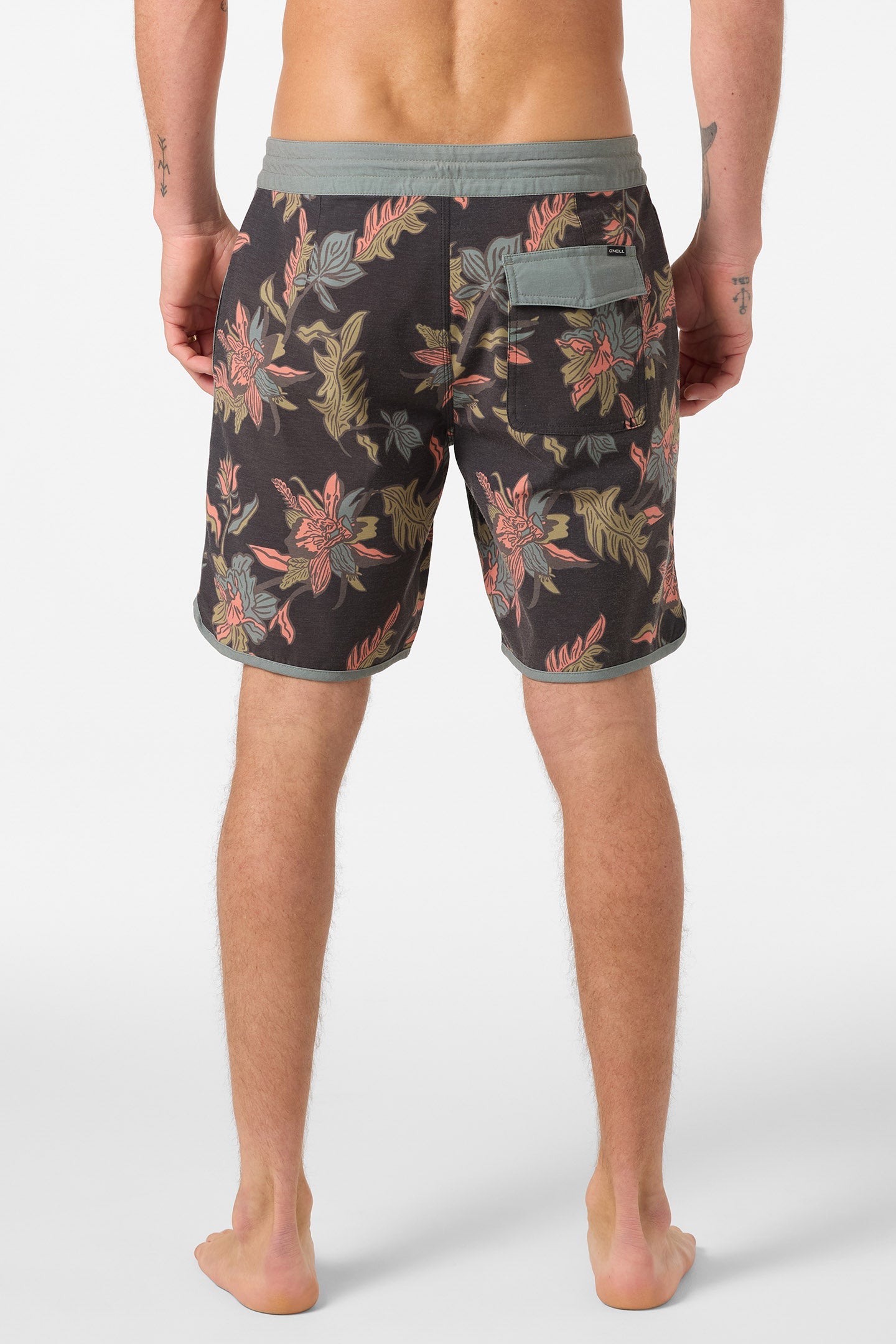 A man stands barefoot with his back to the camera, wearing O'Neill Cruzer Scallop 18 In Boardshorts featuring a colorful floral pattern and a right-side pocket. His relaxed arms display small tattoos on both forearms.