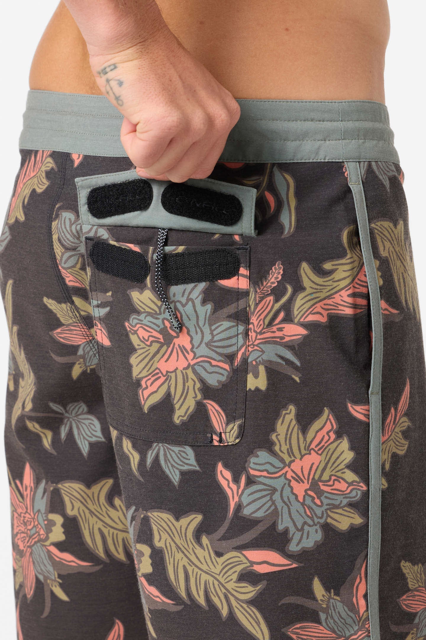A person opens a Velcro back pocket on O'Neill's Cruzer Scallop 18 In Boardshort, featuring a gray waistband, colorful botanical print, and a hidden zippered compartment—all designed for comfort and style.