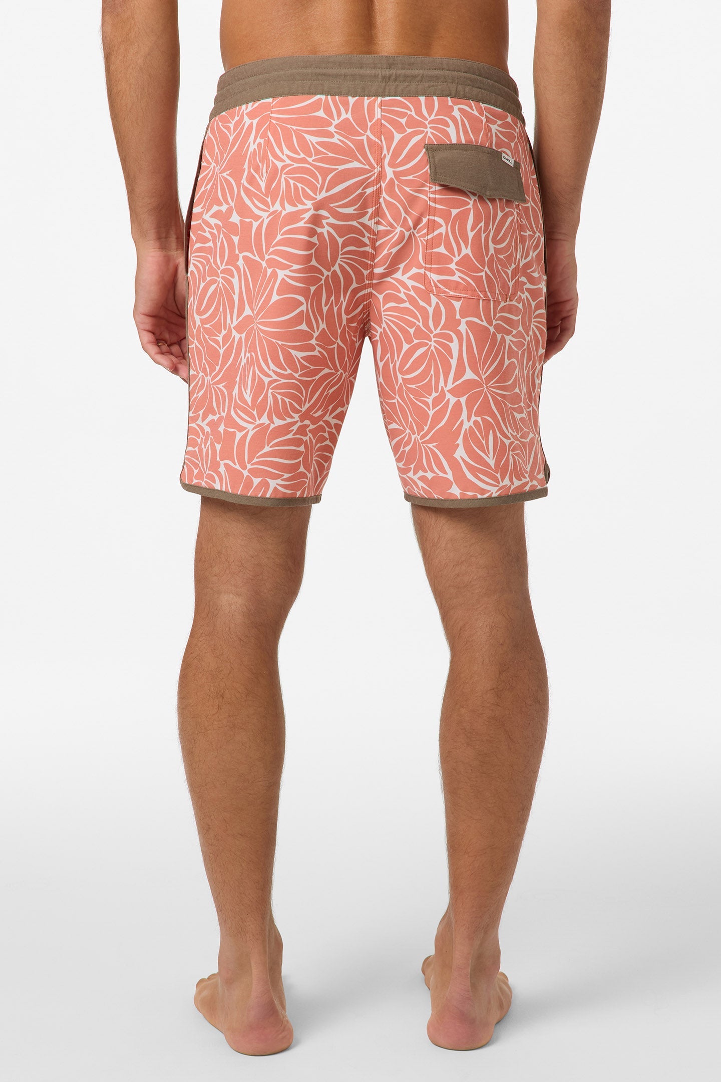 A man stands barefoot, facing away, wearing O'Neill's Cruzer Scallop 18 In Boardshorts—peach with a white leaf print and brown trim. These anti-rash shorts include a back pocket with a buttoned flap. The background is plain white.