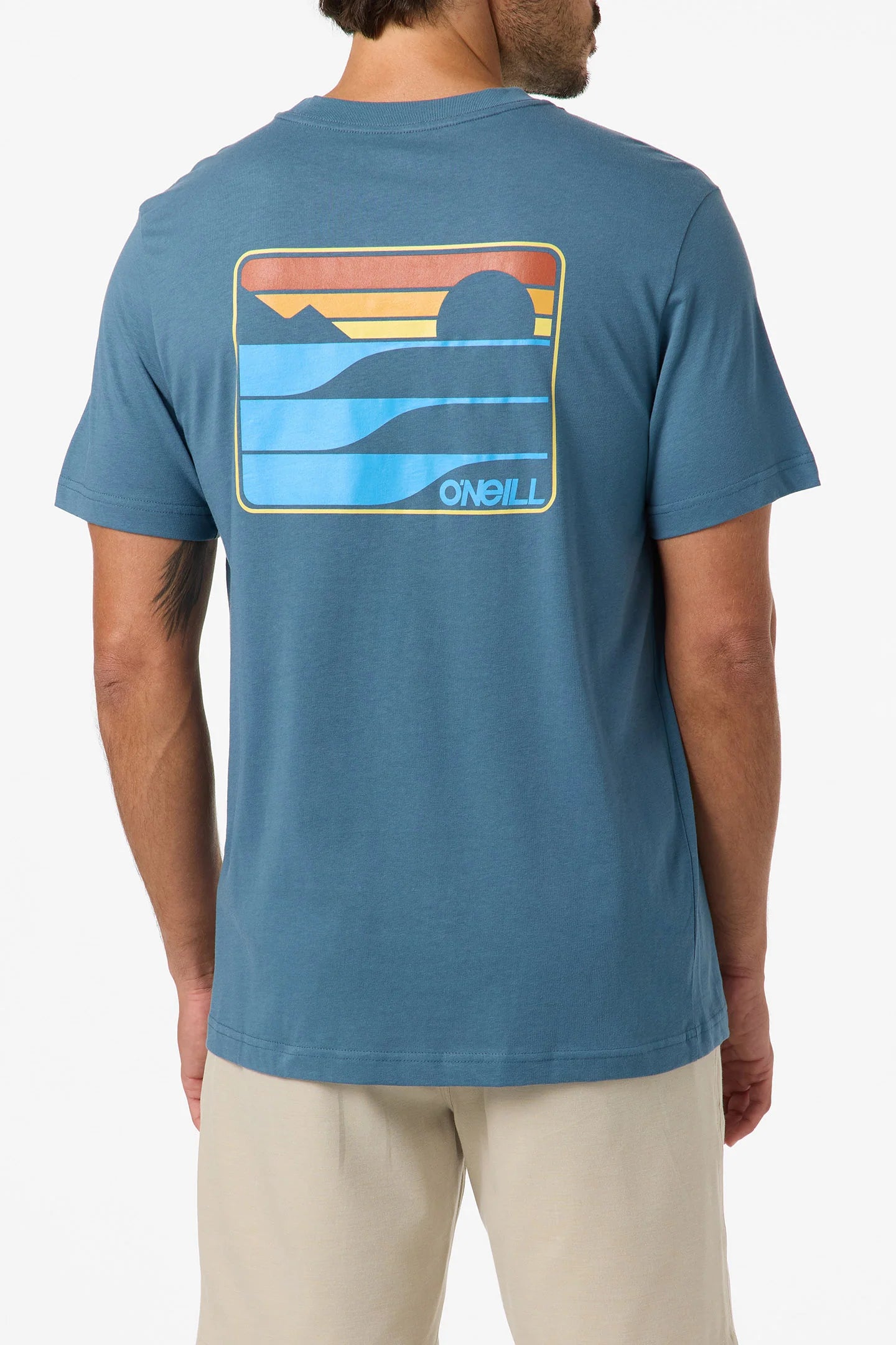 A man wears the O'Neill Dawn Classic Short Sleeve Tee—a blue 100% cotton shirt with a soft hand screen print of a colorful retro sunset, mountains, and waves on the back—paired with light-colored shorts.