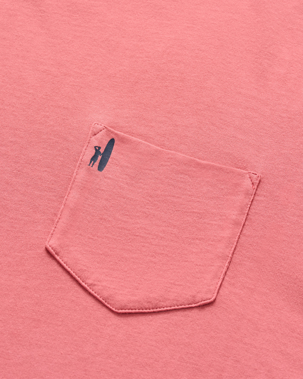 A close-up of the Johnnie-O American Dream pink short sleeve tee's pocket, featuring a small black surfer dude logo—a silhouette of a person with a surfboard—printed near the top left.