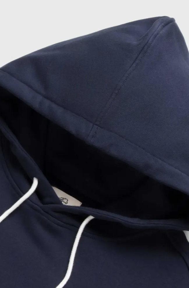 Close-up of the Marine Long Sleeve French Terry Hoodie by Surfside Supply Co., in navy blue, crafted from super soft cotton with a drawstring hood and white ties, laid flat on a light background.