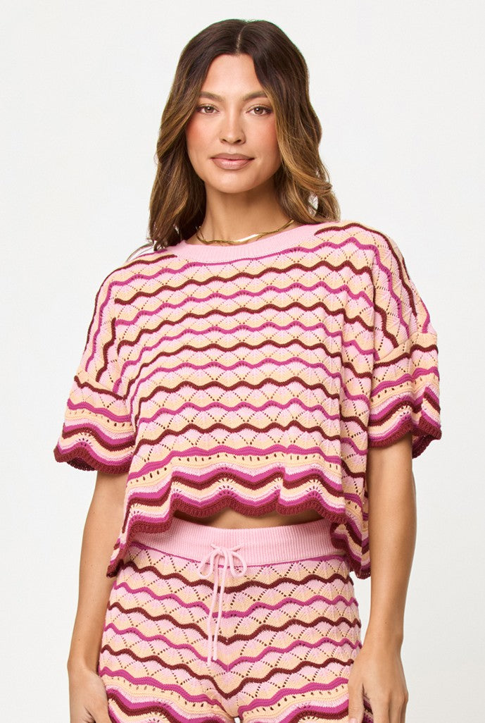 A woman wears the L*Space Berry Spritz Sunchaser Top in a pink and beige zigzag crochet knit with a scalloped hem, styled with matching shorts. She stands against a light background, smiling slightly at the camera.