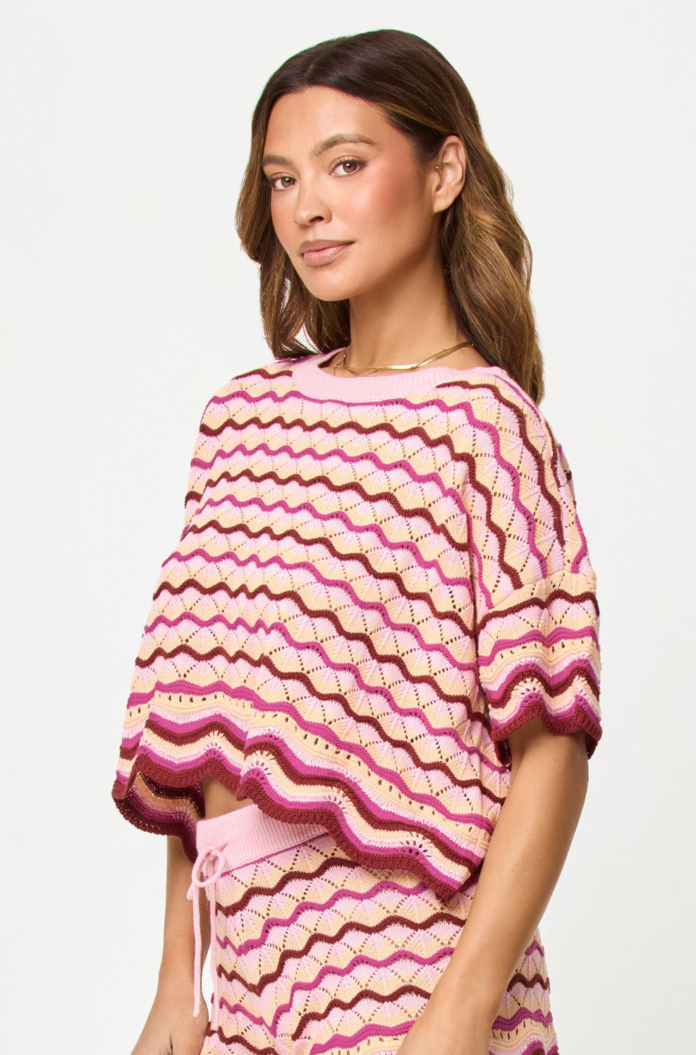 Woman with brown hair wears the L*Space Berry Spritz Sunchaser Top—a pink, red, and cream zigzag crochet knit with a scalloped hem—paired with light pink pants, standing against a plain white background and looking slightly to the side.