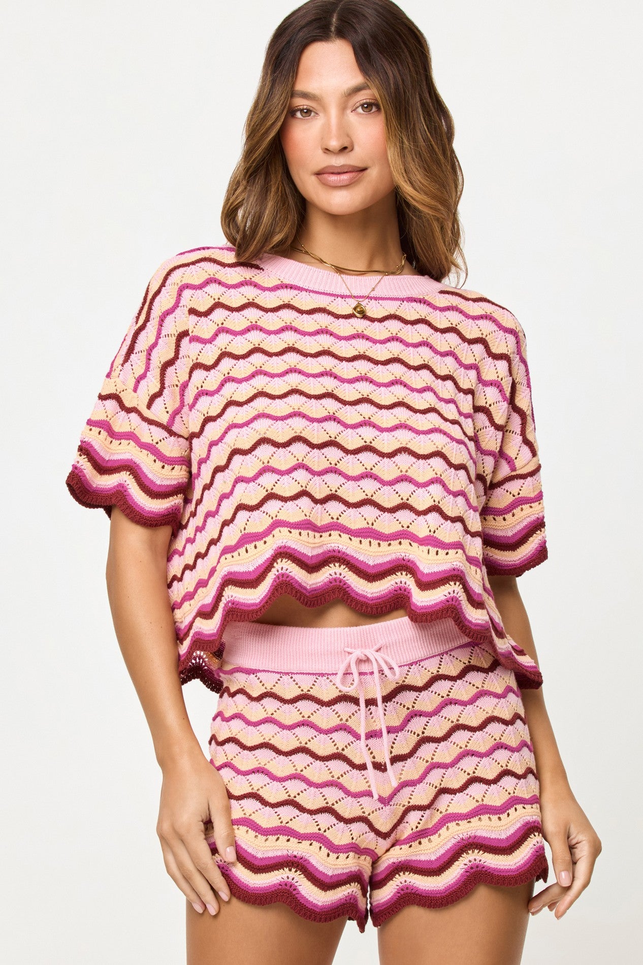 A woman models the L*Space Berry Spritz Sunchaser Top—a pink, purple, and beige wavy striped crochet knit with scalloped hems—paired with matching drawstring shorts against a plain light background.