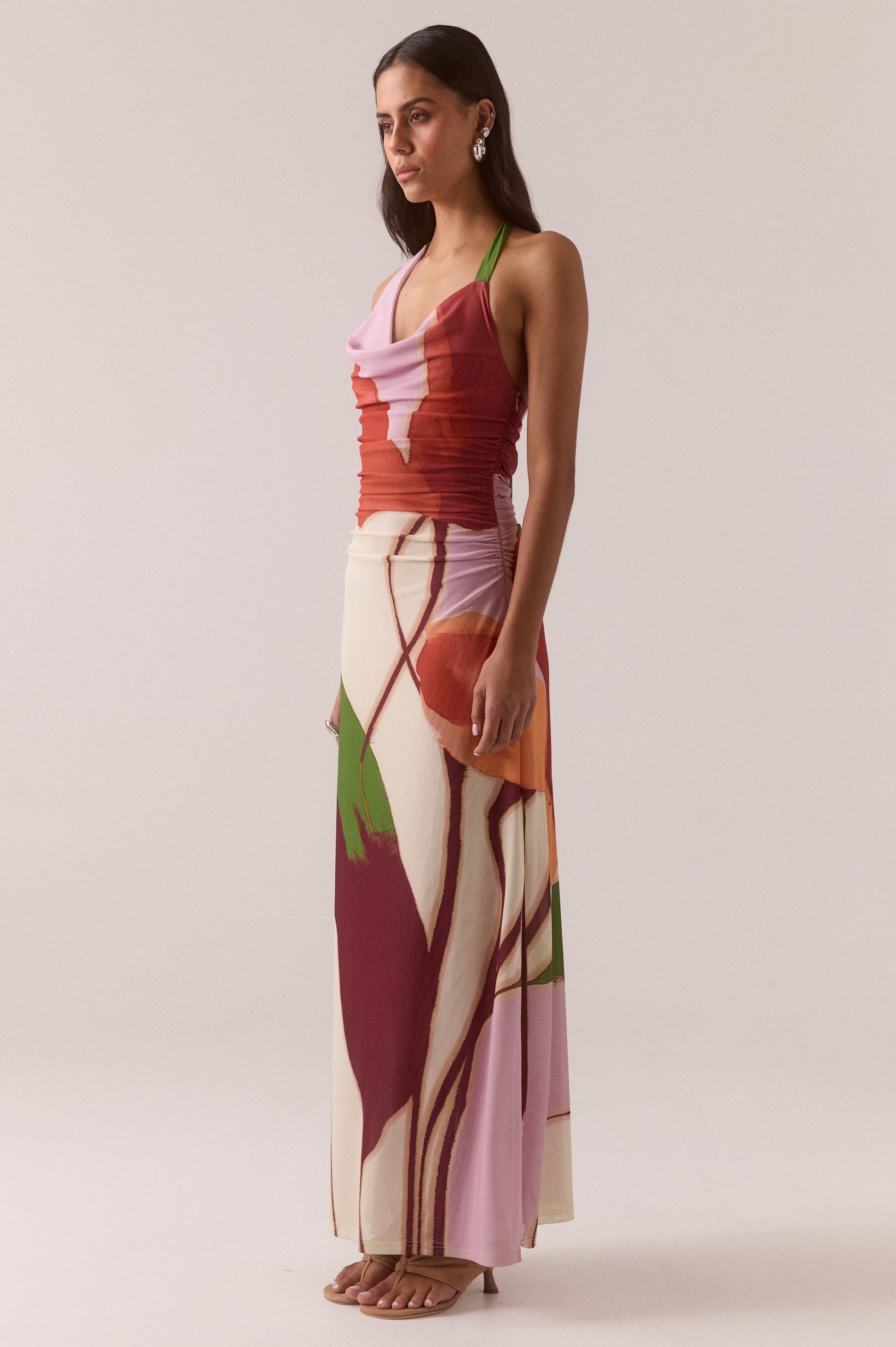 A woman in profile wears the Sovere Kalea Dress, an ankle-length sleeveless stretch mesh style with a colorful abstract floral print in red, pink, cream, and green. She has long dark hair and accessorizes with sandals and earrings.