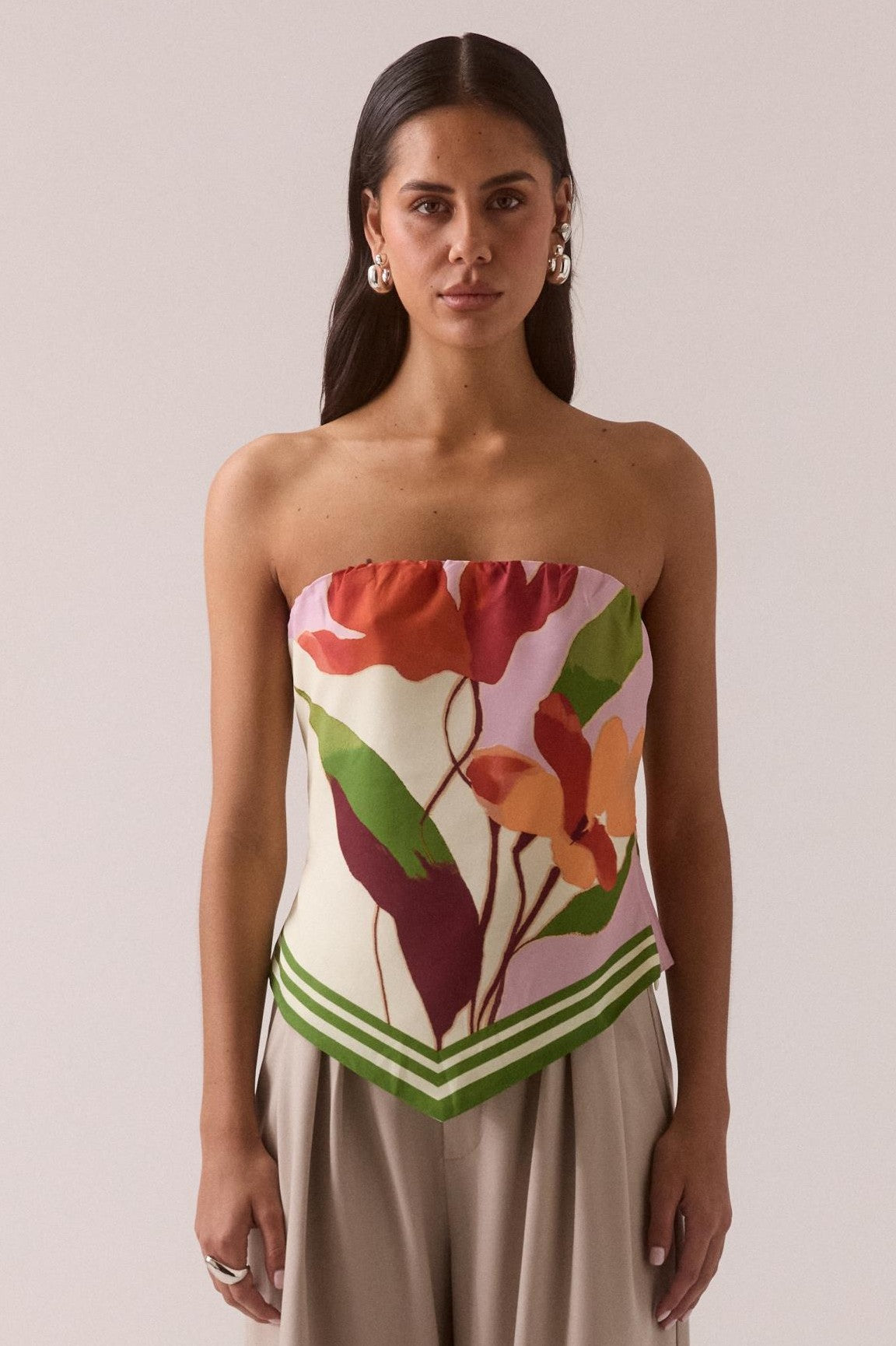A woman with long dark hair wears the Sovere Kalea Top in Ivory Multi, a strapless design with an exclusive print and green-striped hem, paired with wide beige pants. She stands facing forward against a neutral background.
