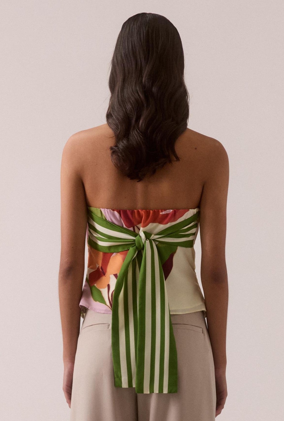 A woman with wavy dark hair faces away, wearing Sovere's Kalea Top—a strapless crepe piece with an exclusive placement print and a striking green and white striped bow at the back—paired with light beige pants.