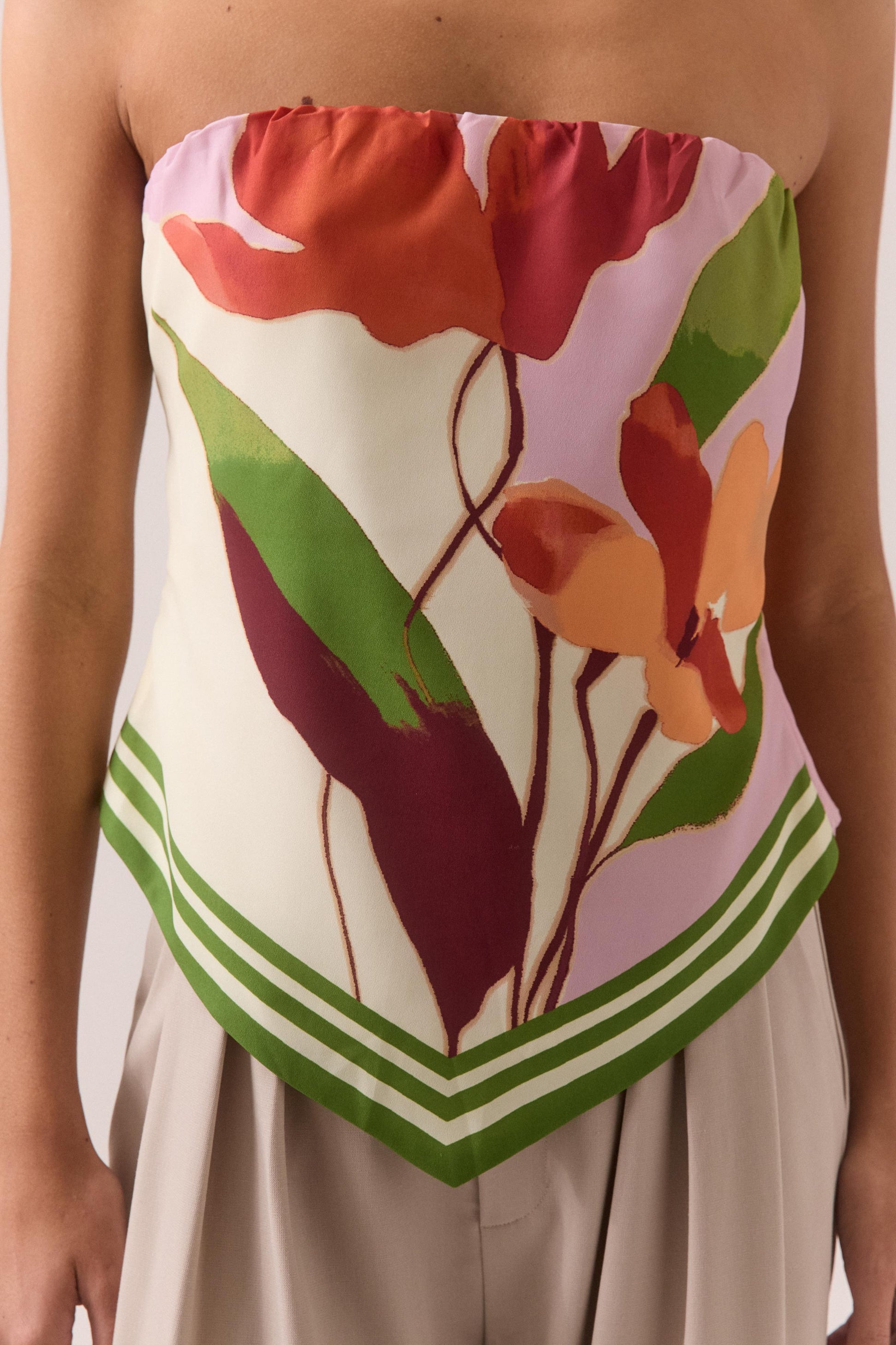 A person wears the Sovere Kalea Top, a strapless crepe piece with an exclusive placement print in bold red, orange, and green tones plus a green-striped hem, paired stylishly with beige high-waisted pants.