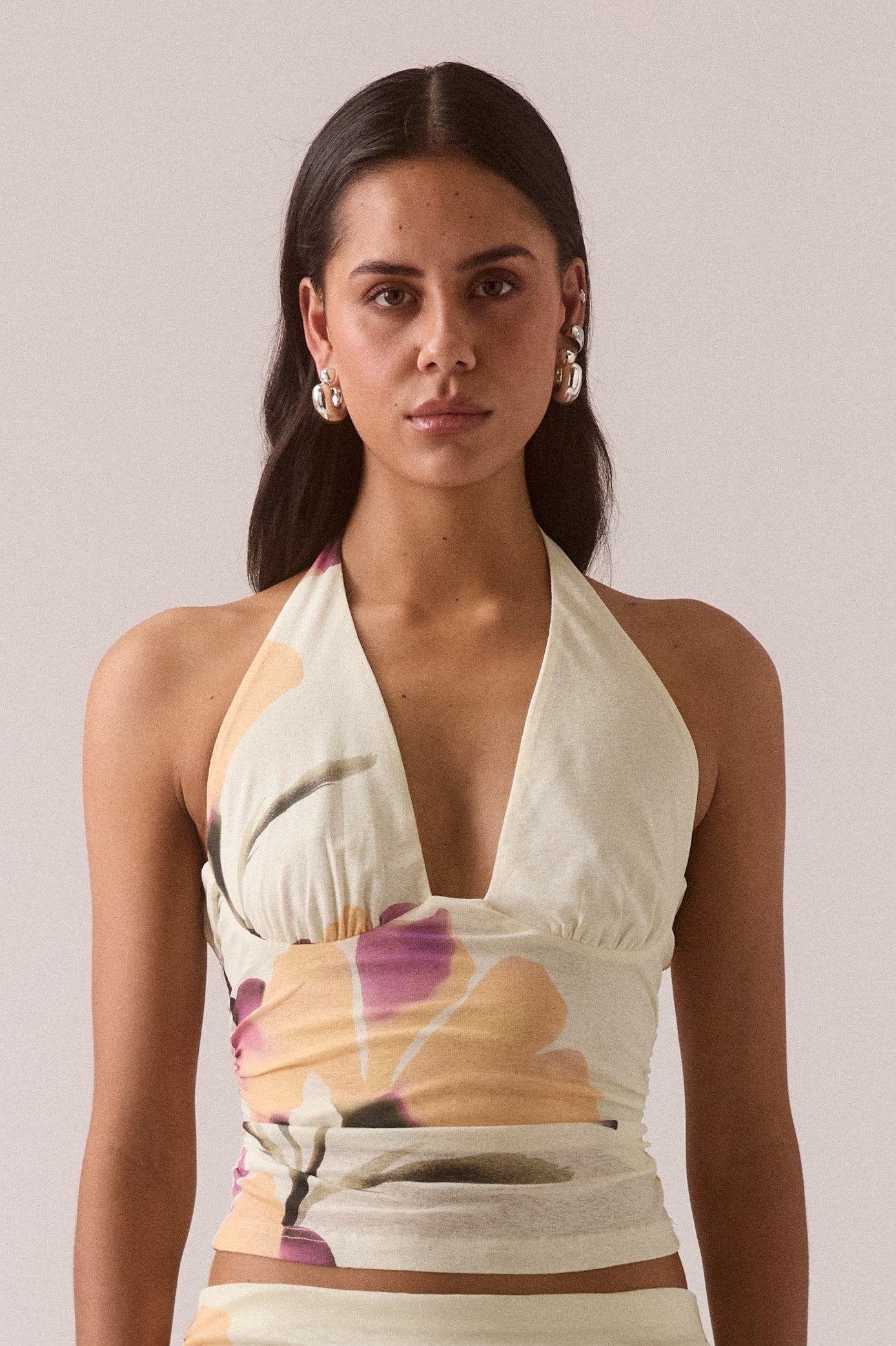 A woman with long dark hair wears the Sovere Akanni Top in Yellow Multi, a sleeveless halter neck made from stretch mesh with an exclusive abstract floral print. She pairs it with silver hoop earrings and has a neutral expression.