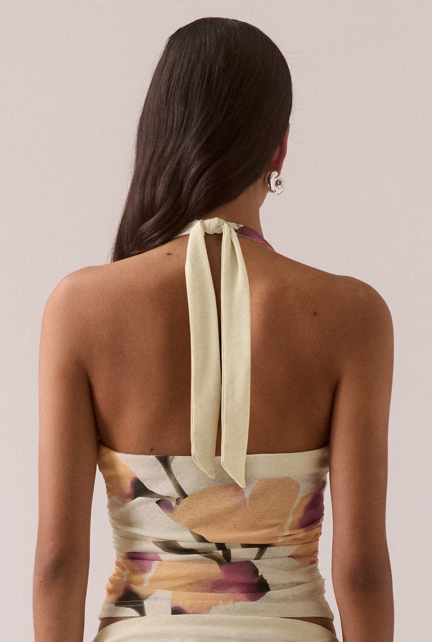 A woman with long, dark hair is seen from the back wearing the Sovere Akanni Top, a fitted women's halter neck style with a large yellow, purple, and beige floral print and wide fabric straps that tie at the neck.