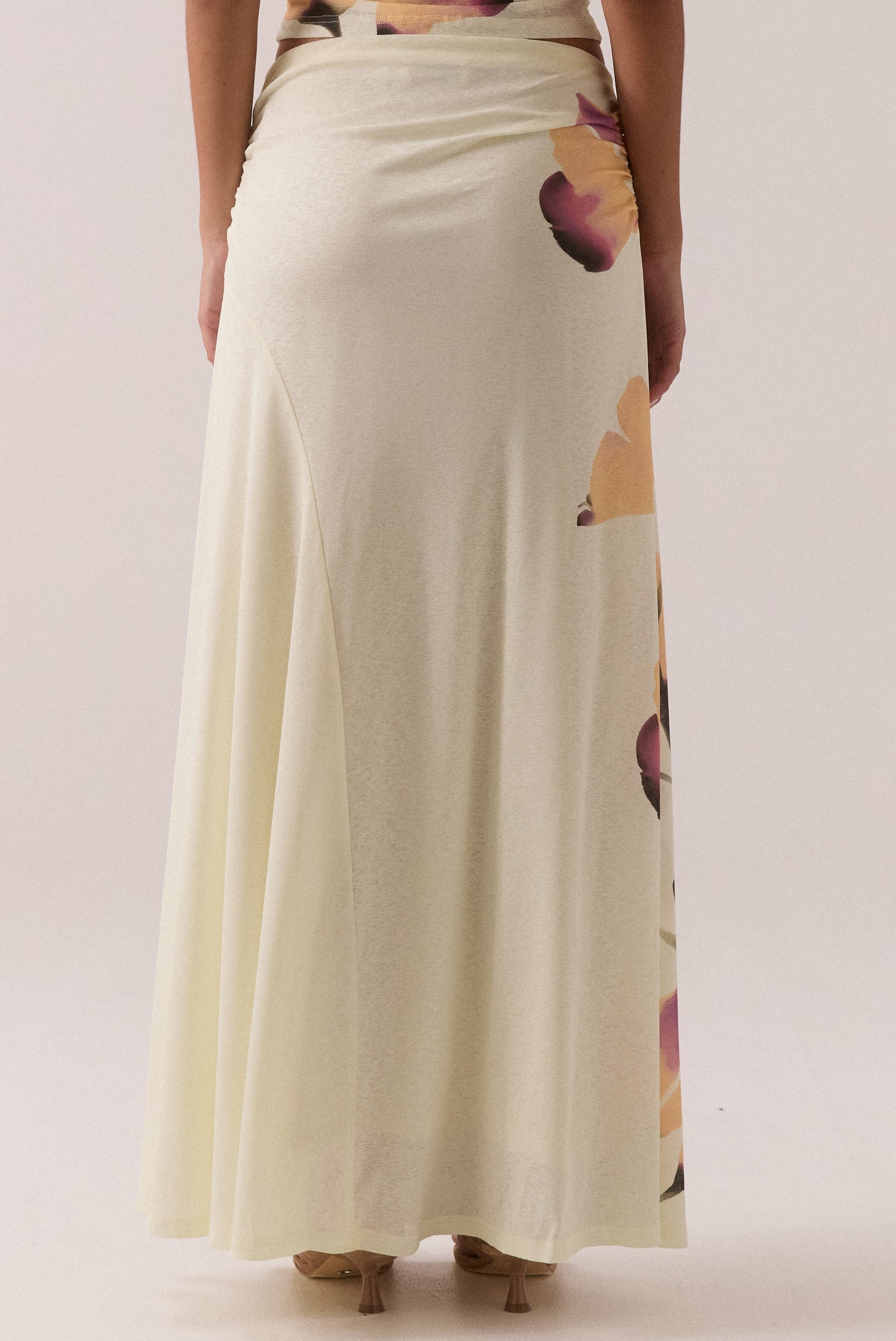 A person stands with their back to the camera against a plain background, wearing Sovere's Akanni Skirt—a long, cream-colored mid-rise piece featuring a subtle floral pattern.