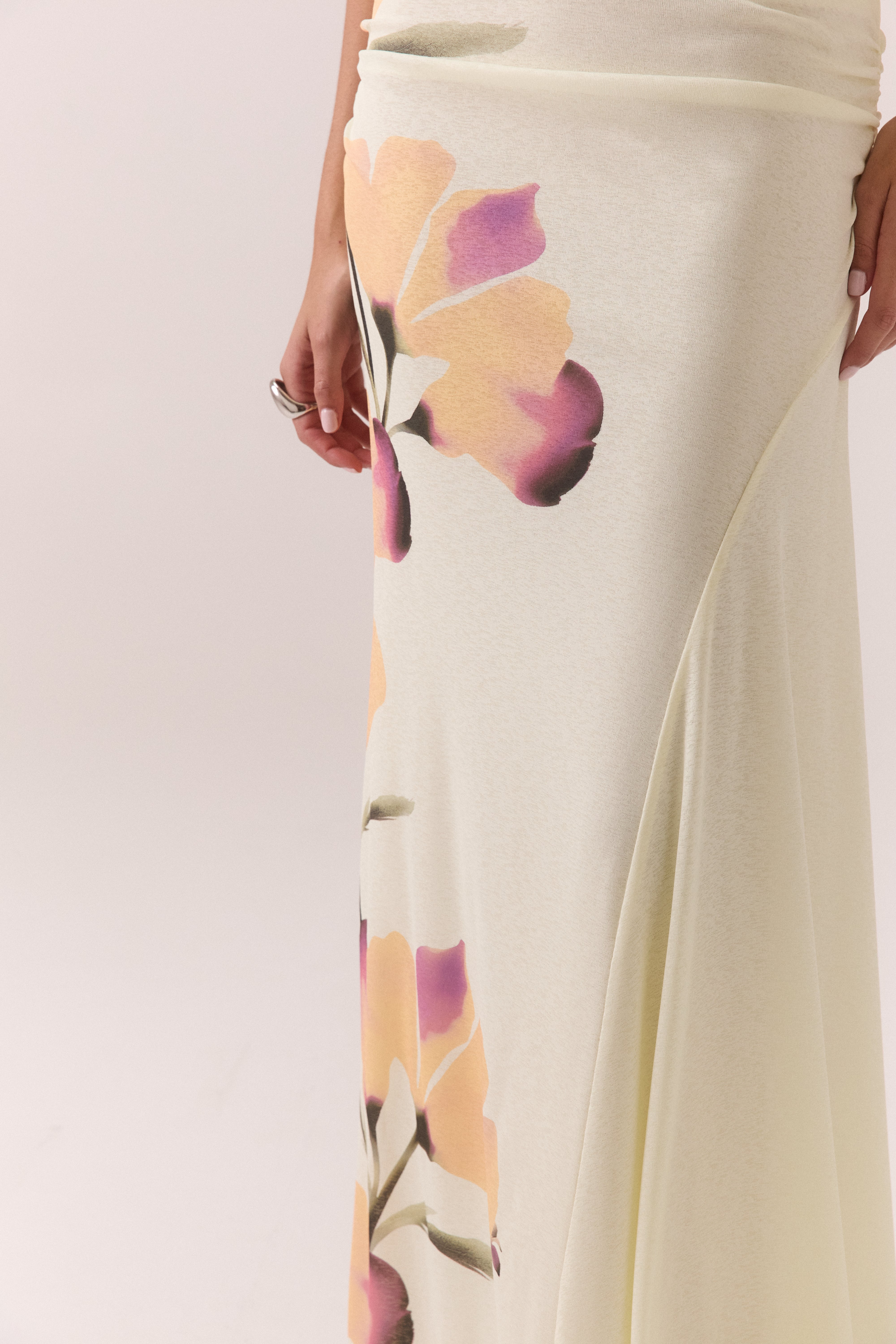 A person wears the Akanni Skirt by Sovere, a light cream mid-rise skirt featuring large abstract floral patterns in pink, purple, and yellow. Only part of the torso and skirt are visible, with one hand resting at their side.