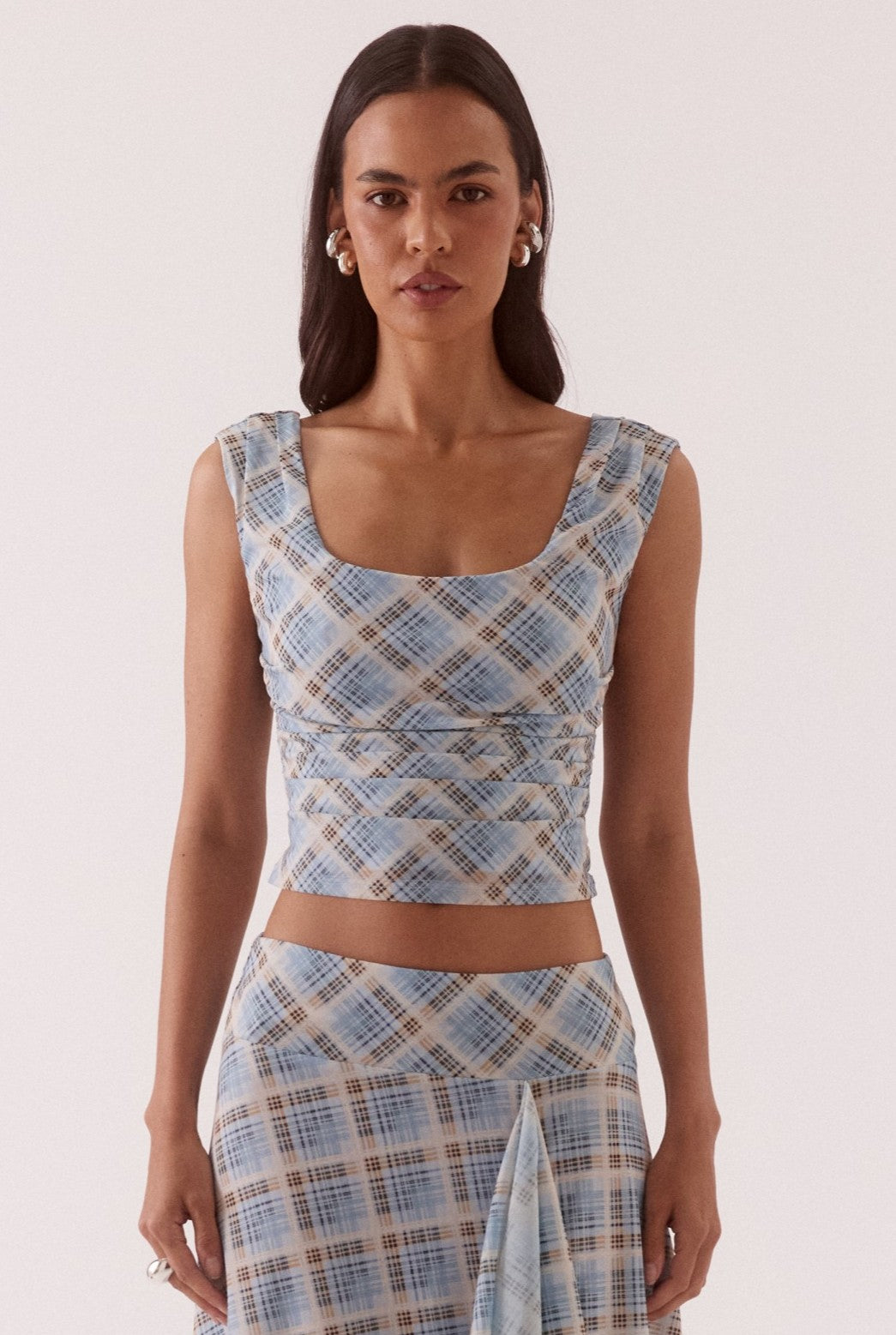 A woman wears the Sovere Novalyn Plaid Top in Blue Multi, styled with a matching high-waisted skirt. She has long straight hair and completes her look with statement earrings and rings.