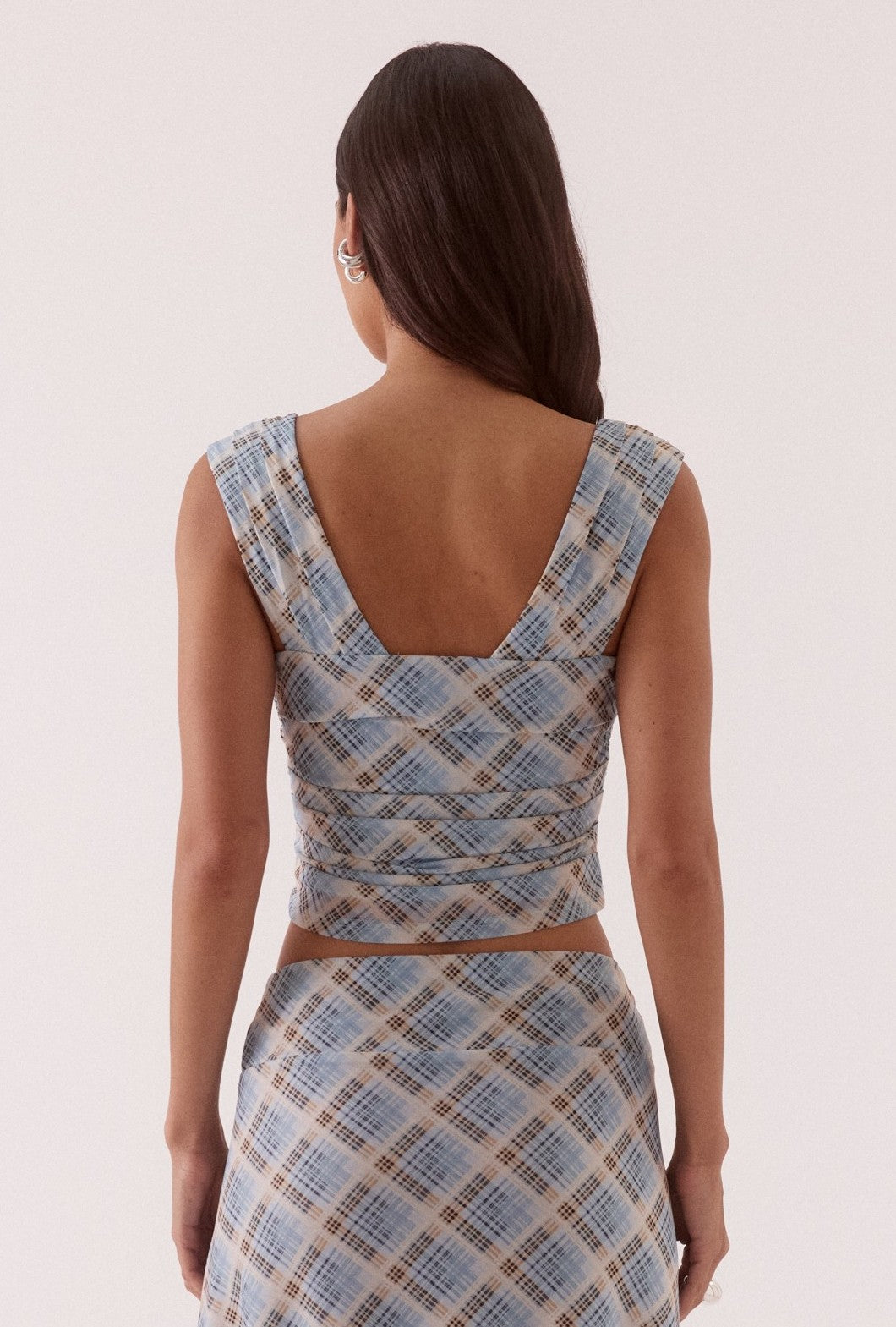 A woman with long dark hair faces away, wearing the Novalyn Plaid Top by Sovere with a matching high-waisted skirt in blue and beige plaid. A light background emphasizes the clean lines of the outfit.