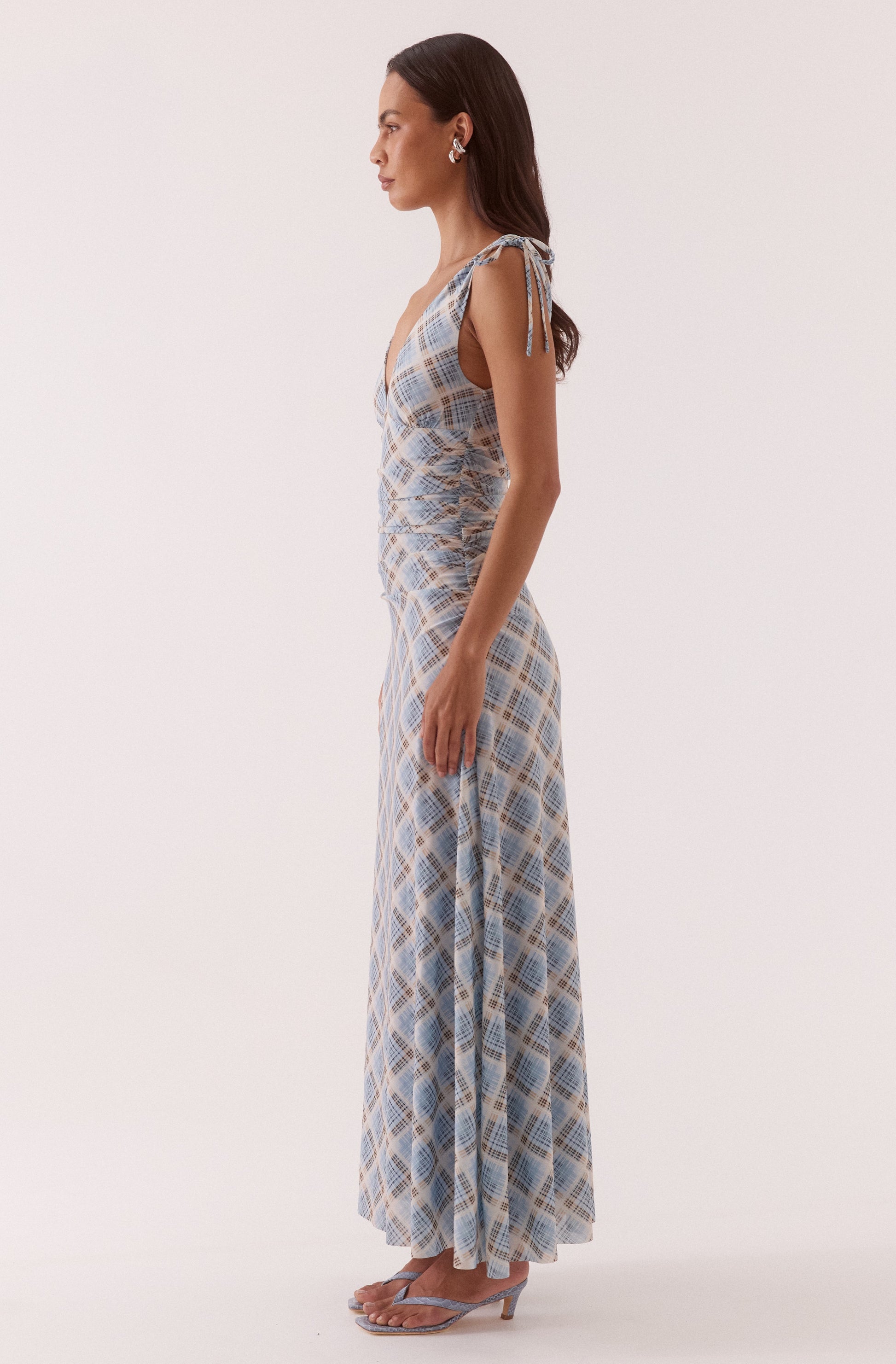 A woman stands in profile wearing the Sovere Novalyn Plaid Sleeveless Dress, featuring blue and white plaid, ruched sides, tie straps, and clear-heeled sandals against a plain light background.