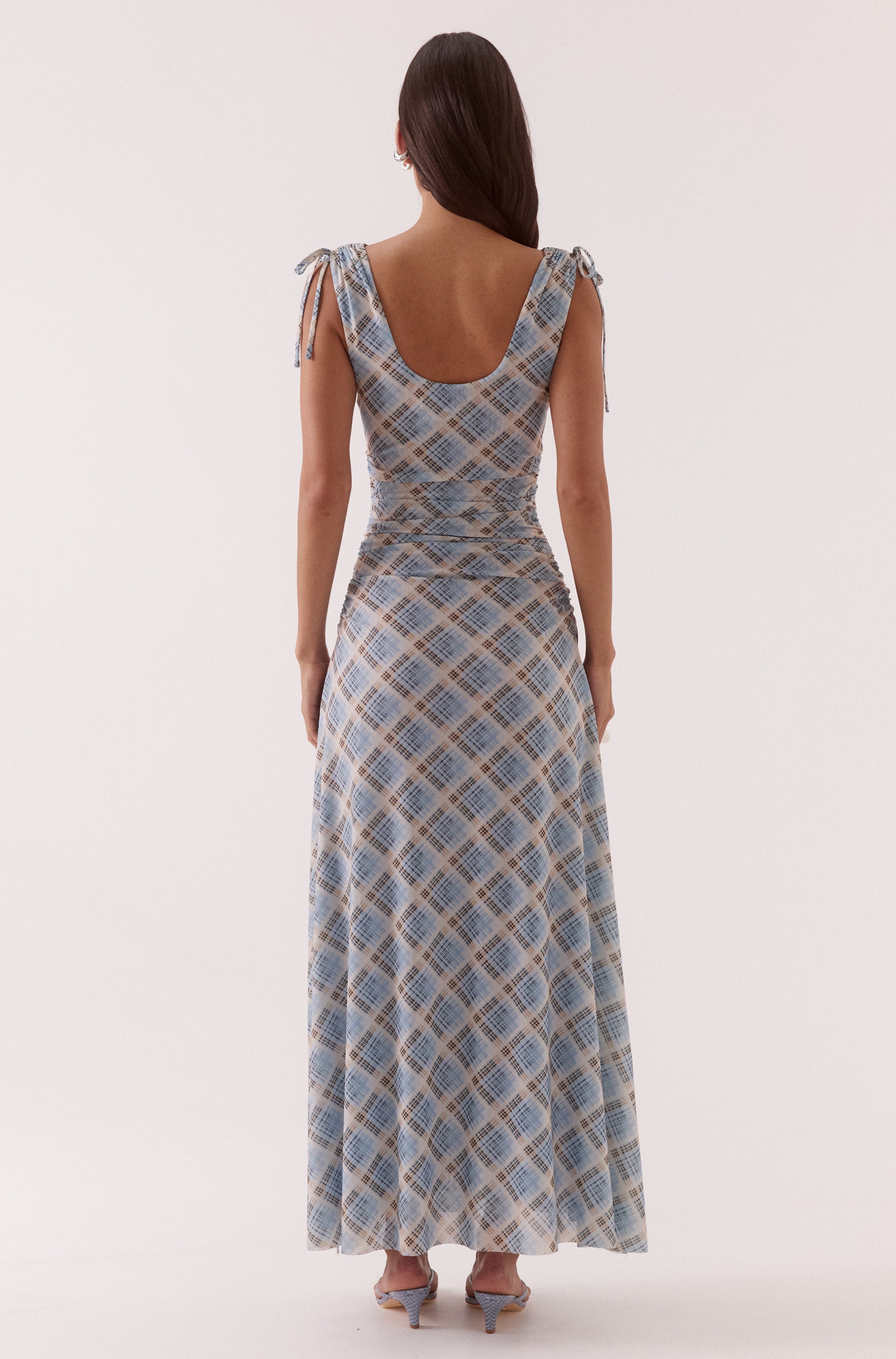 A woman with long brown hair faces away, wearing the Sovere Novalyn Plaid Sleeveless Dress in blue and beige with tied shoulder straps, scoop back, and heeled sandals.