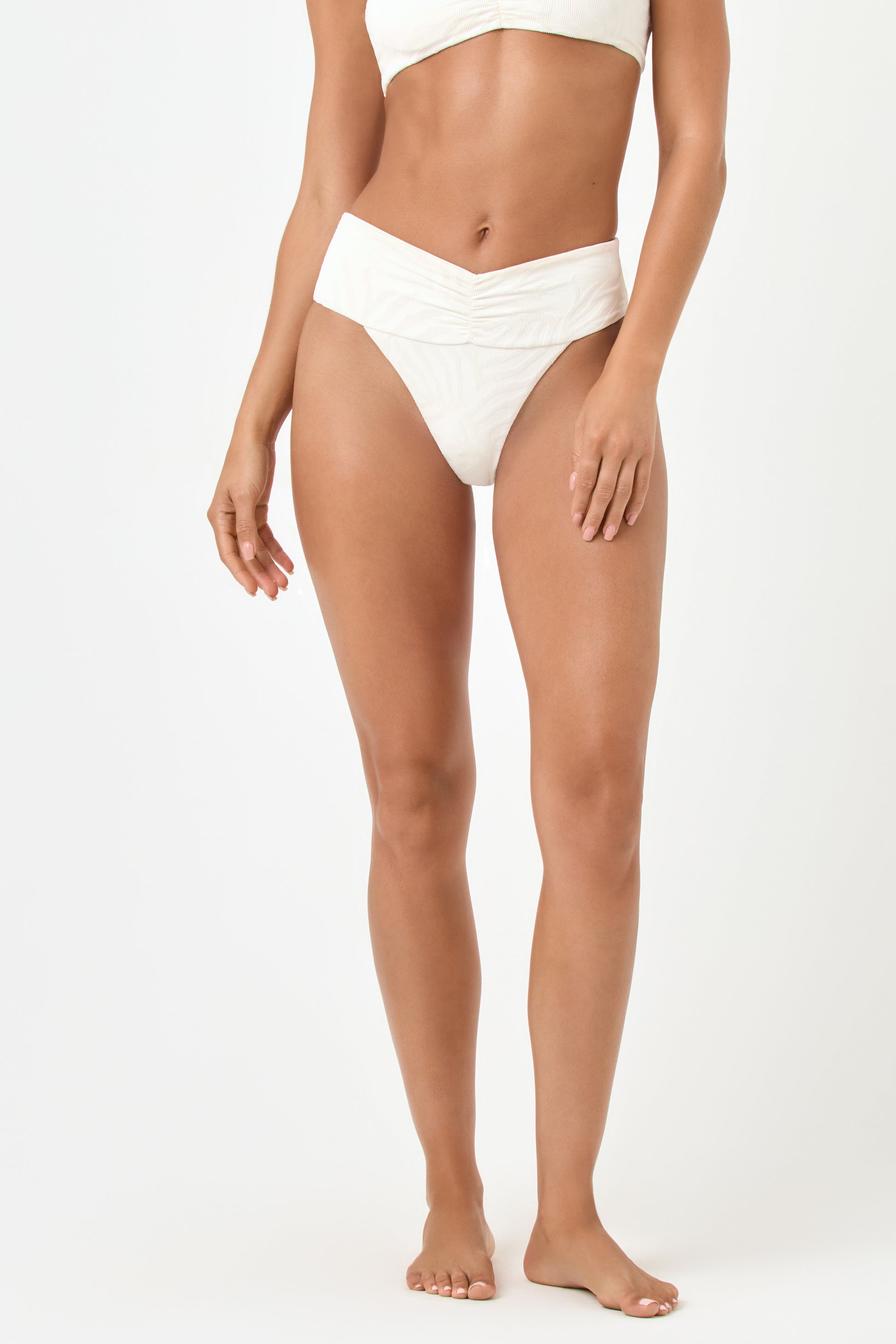 A woman stands barefoot against a white background, wearing the cream Sail Along Bardot Bottom from LSpace, featuring a subtle textured pattern and matching bikini top. Her arms rest relaxed at her sides.