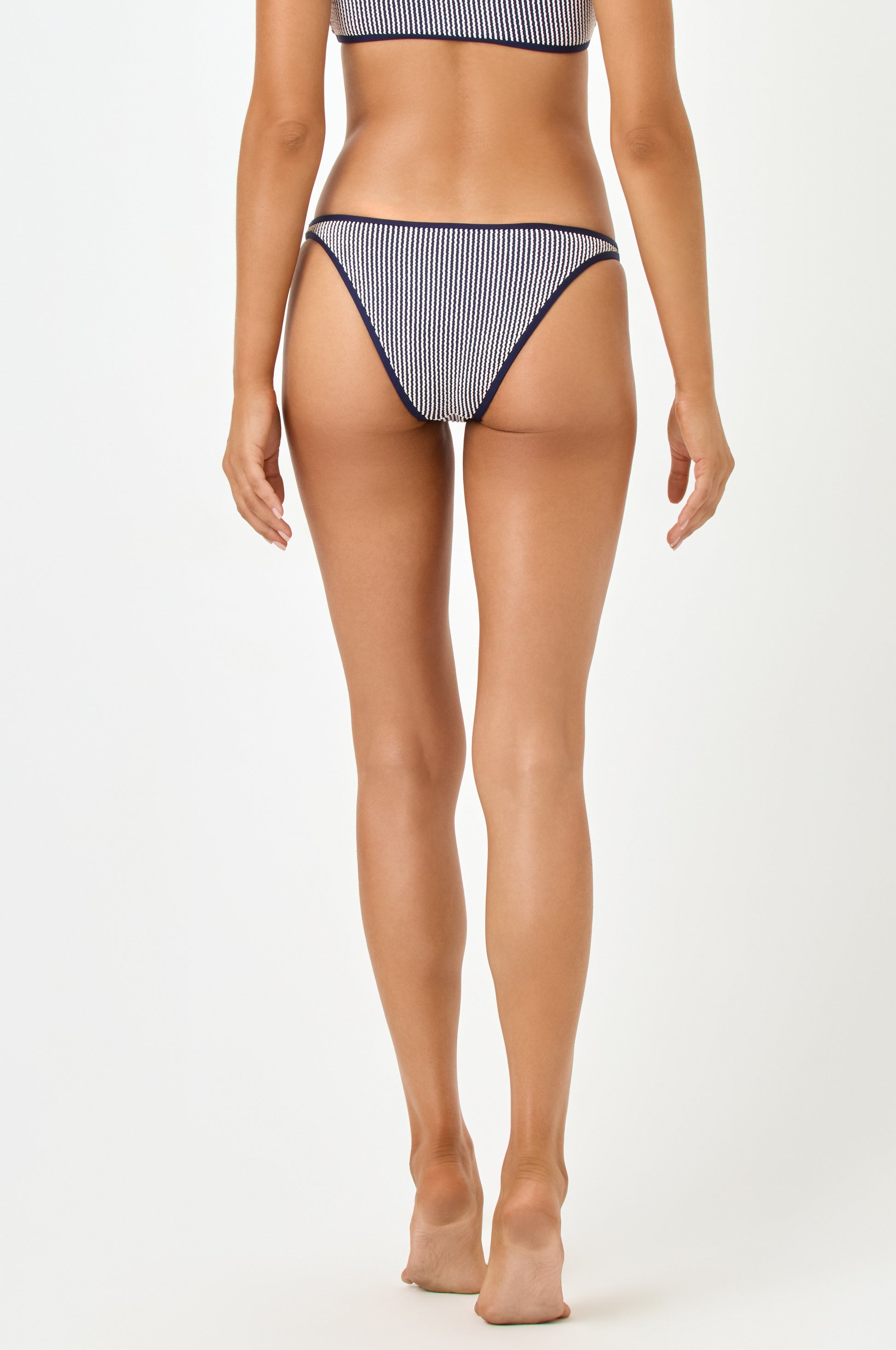 A woman stands barefoot with her back to the camera, wearing the LSpace Midnight Blue Suzi Bottom and matching top in ribbed fabric. The background is plain white.