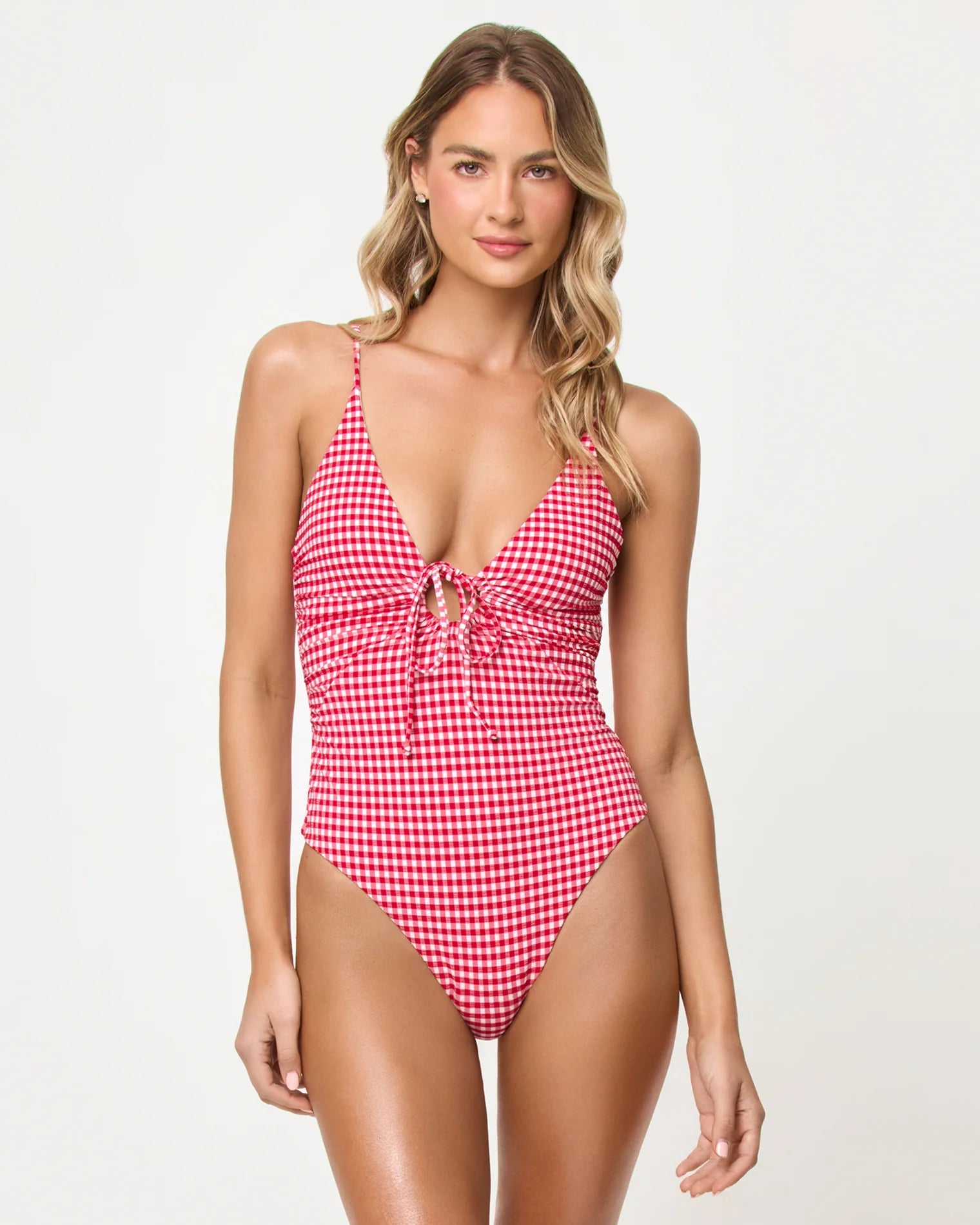 A woman models the Atta Girl Gingham Piper One Piece by LSpace in Red Multi, featuring a front tie, adjustable straps, and a deep V neckline, against a plain light background.