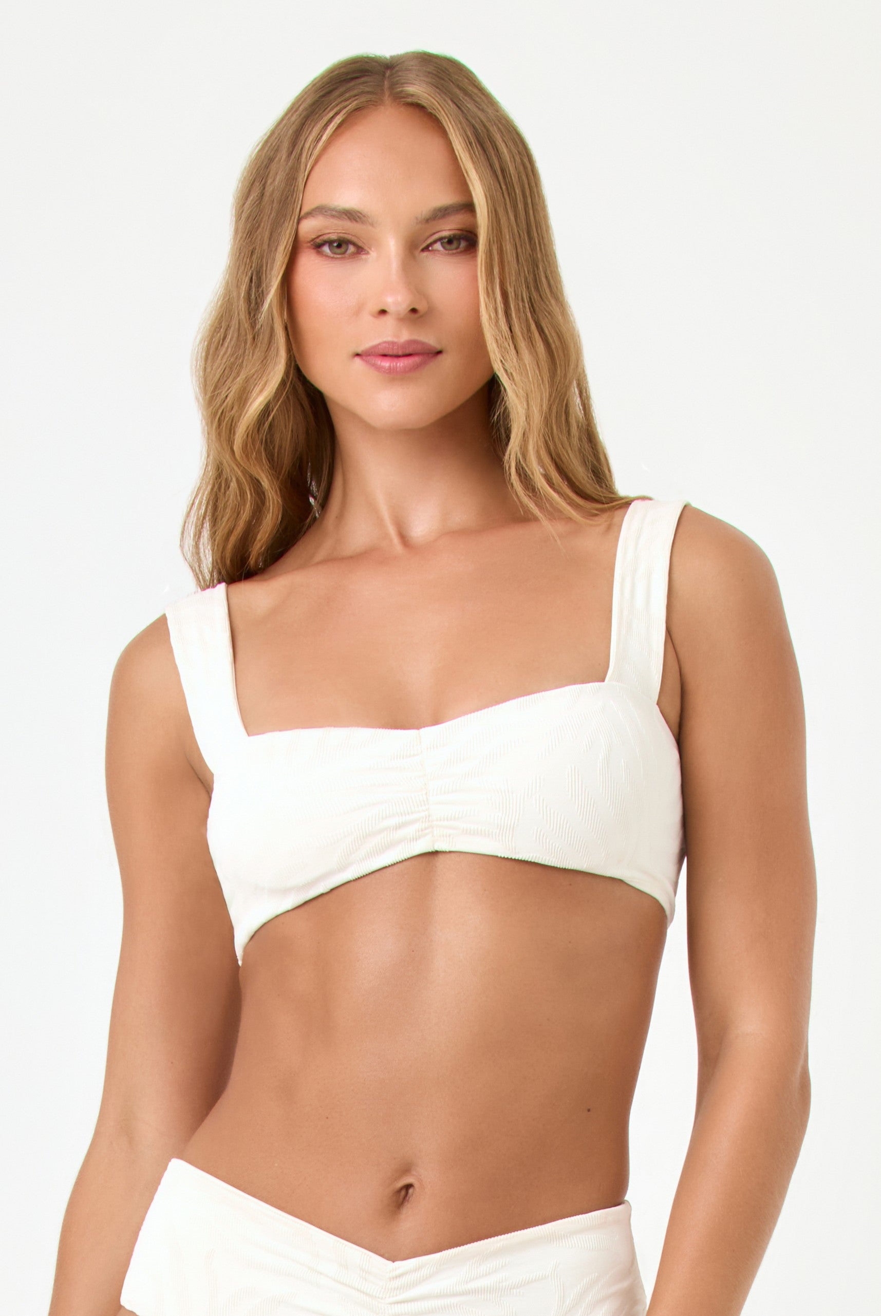A woman with long, wavy blonde hair wears the LSpace Sail Along Stripe Marlee Top, a white bralette-style swimsuit with wide straps, standing against a plain light background and smiling slightly at the camera.