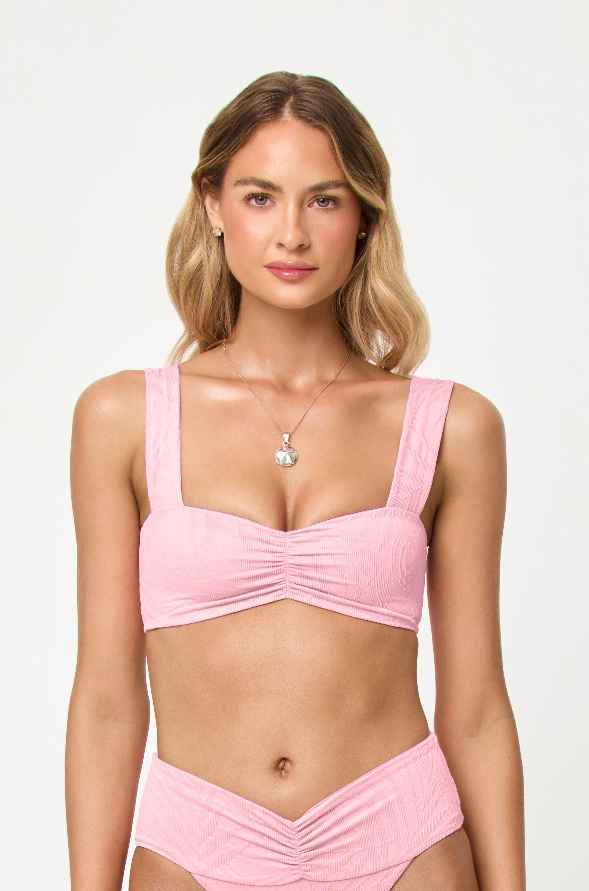 A woman with light brown hair wears the LSpace Pink Sugar Marlee Top bikini in pink, paired with matching high-waisted bottoms and a silver pendant necklace, standing against a plain white background and facing forward.