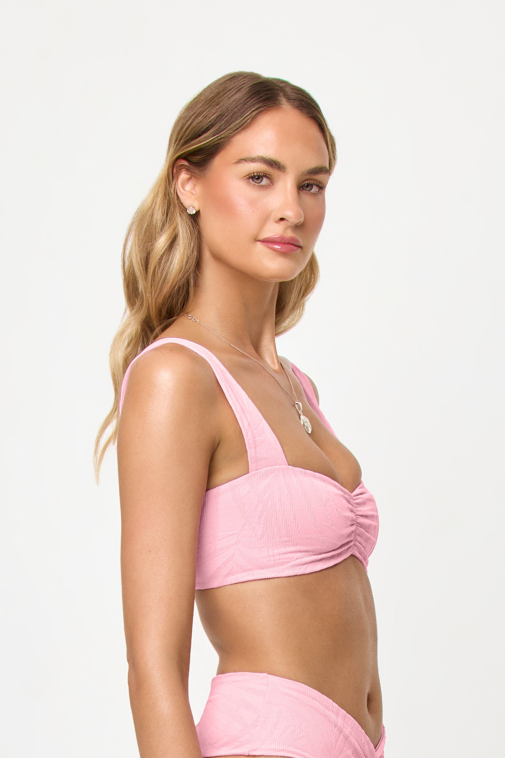 A woman with wavy blonde hair poses confidently in the LSpace Pink Sugar Marlee Top, a light pink bikini featuring front ruching and thick straps, against a plain white background.