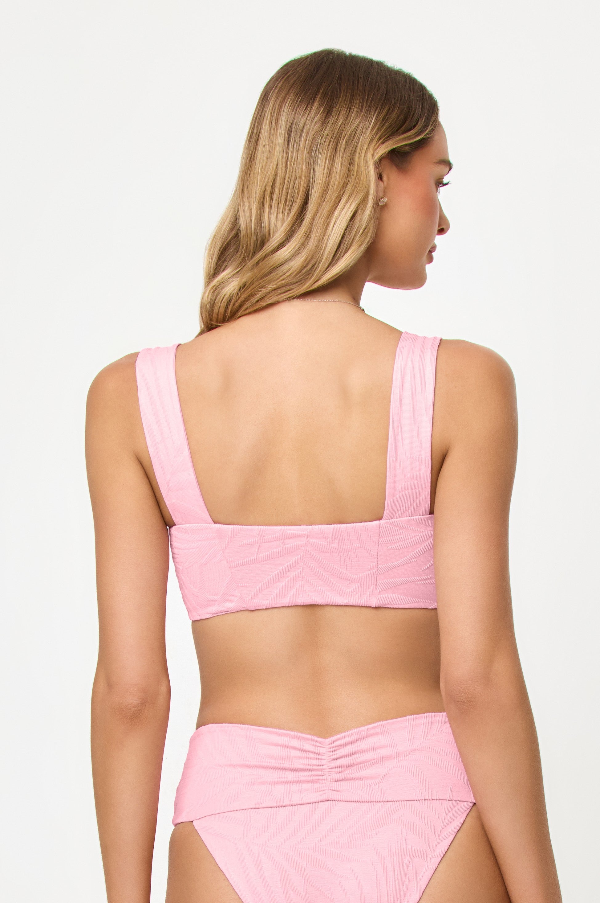 A woman with wavy, light brown hair is seen from the back wearing the LSpace Pink Sugar Marlee Top bikini, featuring thicker straps and high-waisted bottoms, set against a plain white background.