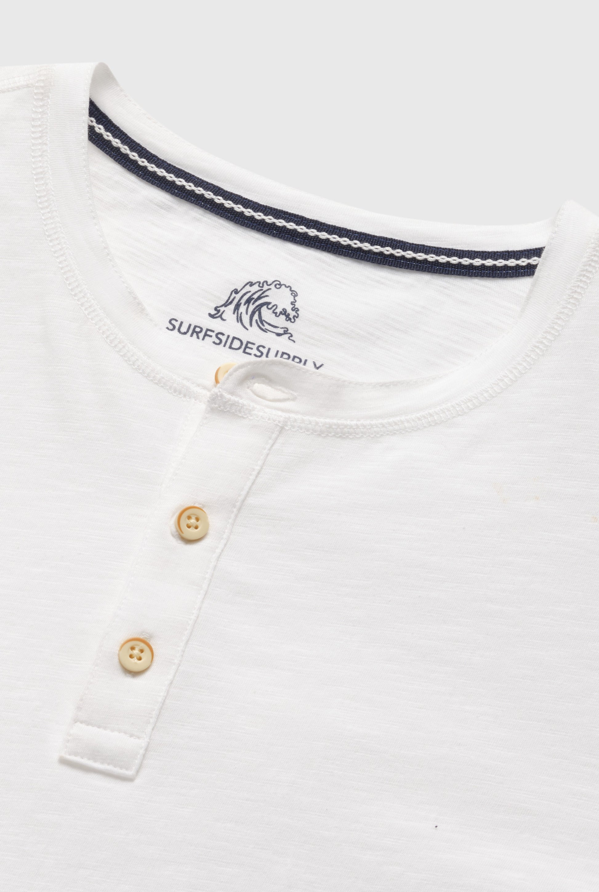 Close-up of the Surfside Supply Co. Sean Short Sleeve Garment Dye Slub Henley, a regular fit cotton shirt with three light brown buttons, navy-striped neckline, and an inner label featuring a wave graphic and “SURFSIDE SUPPLY.”.