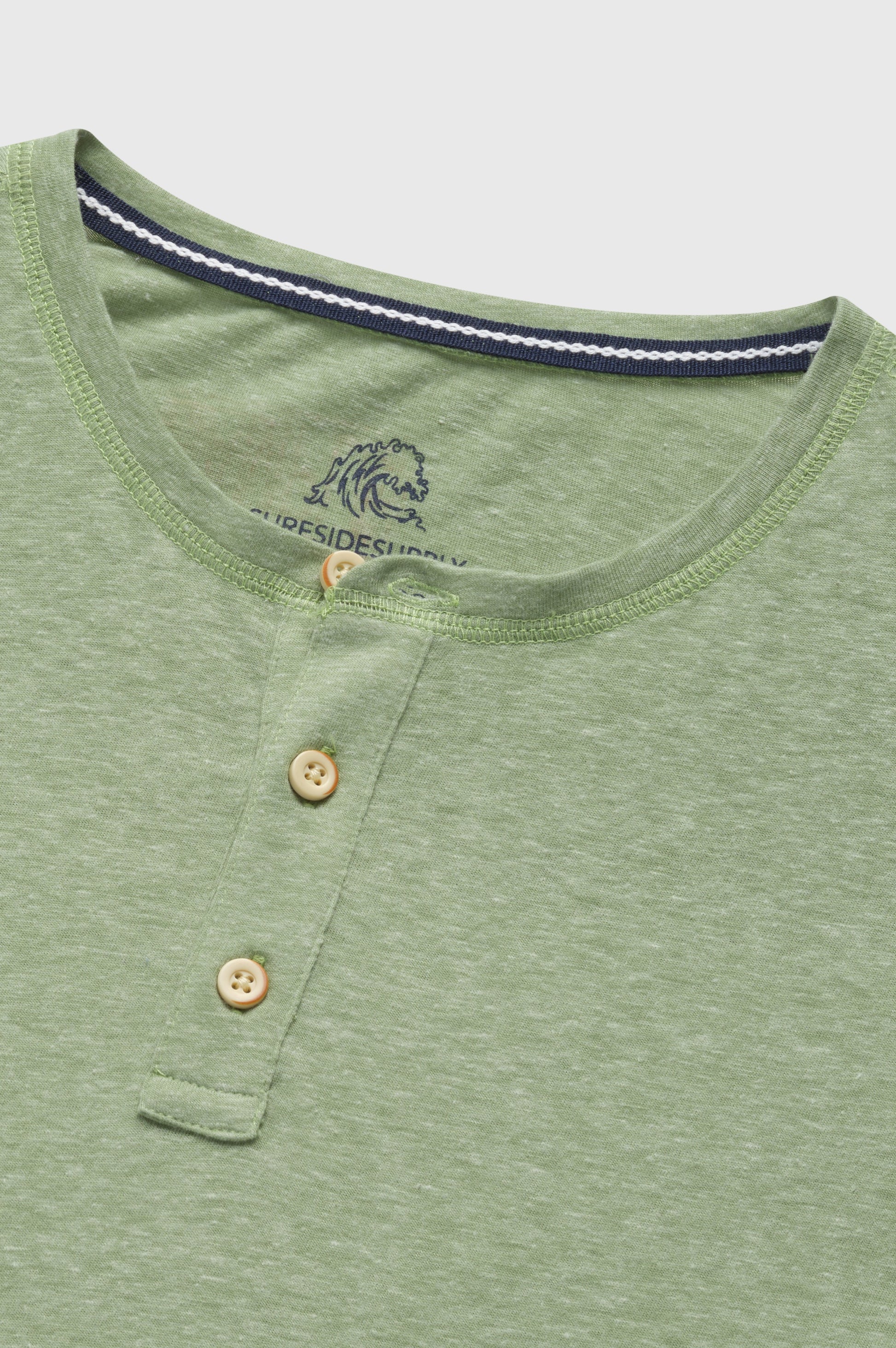 The Sean Short Sleeve Single Dye Jersey Henley by Surfside Supply Co. is a light green shirt with a round neck, two tan buttons on the short placket, and a subtle embroidered logo inside the collar.