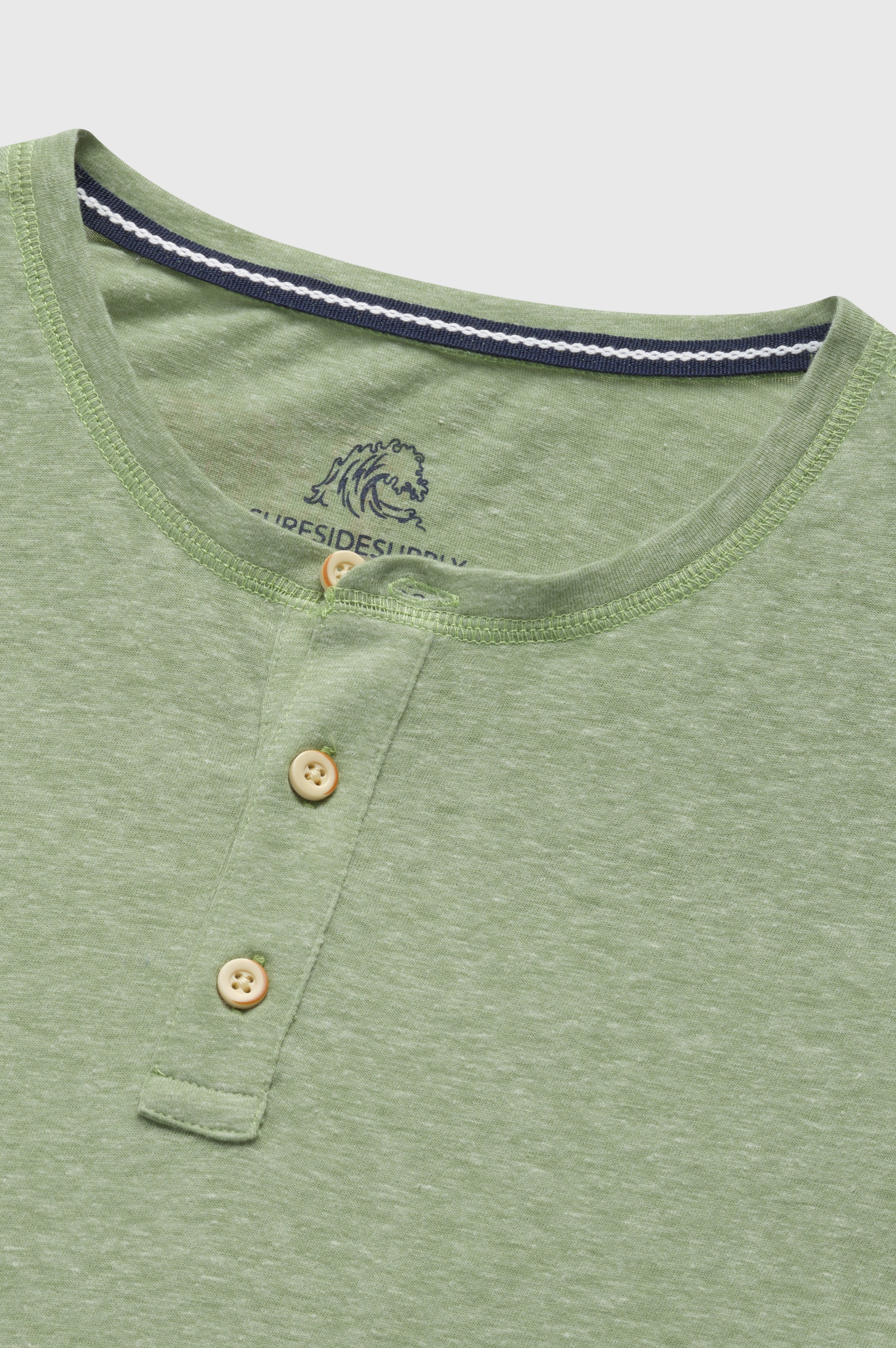The Sean Short Sleeve Single Dye Jersey Henley by Surfside Supply Co. is a light green shirt with a round neck, two tan buttons on the short placket, and a subtle embroidered logo inside the collar.