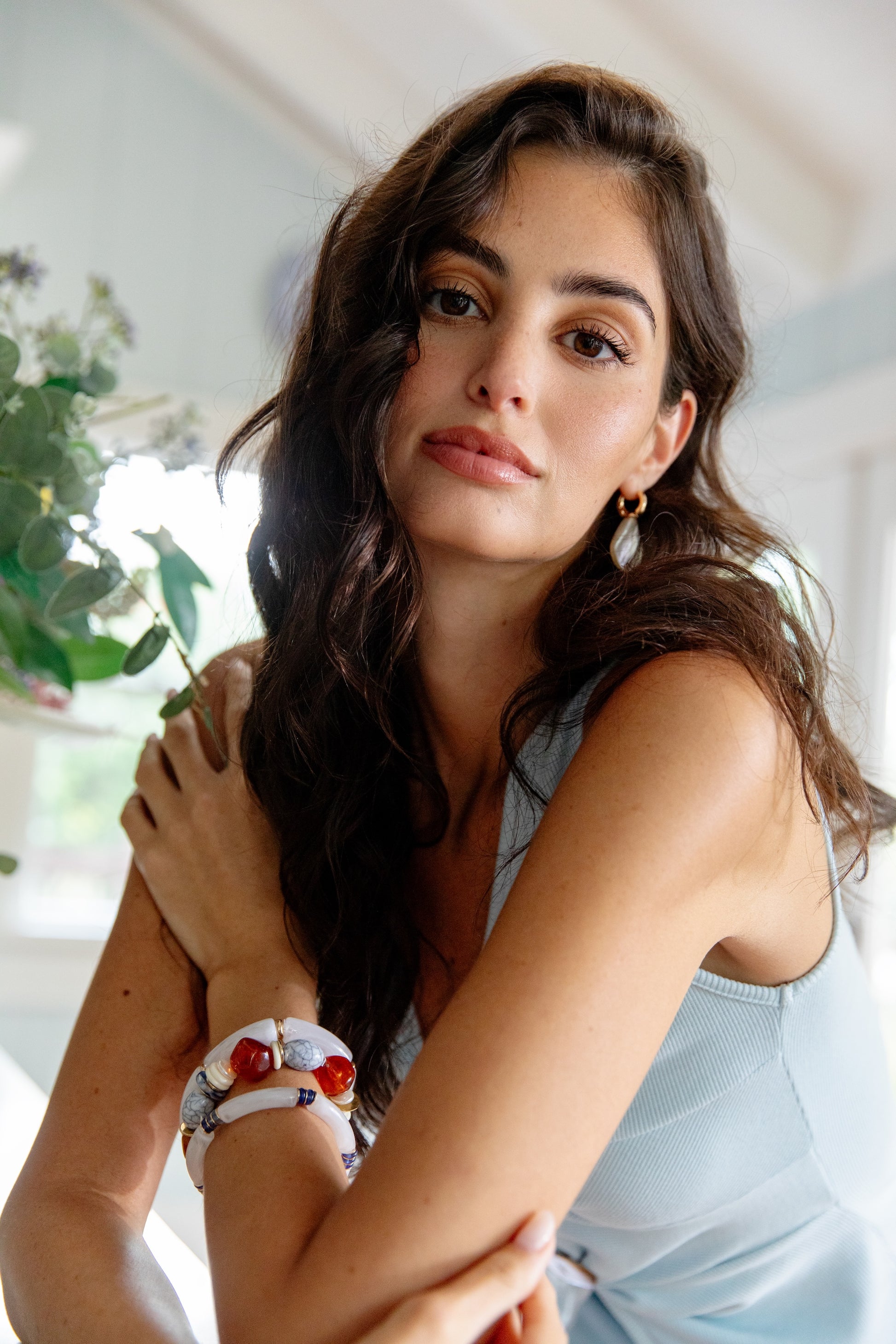 A woman with long dark hair, wearing Sun Kissed Pearl Earrings by Ettika and a light blue sleeveless top, sits indoors near greenery, looking calmly at the camera with a relaxed expression.