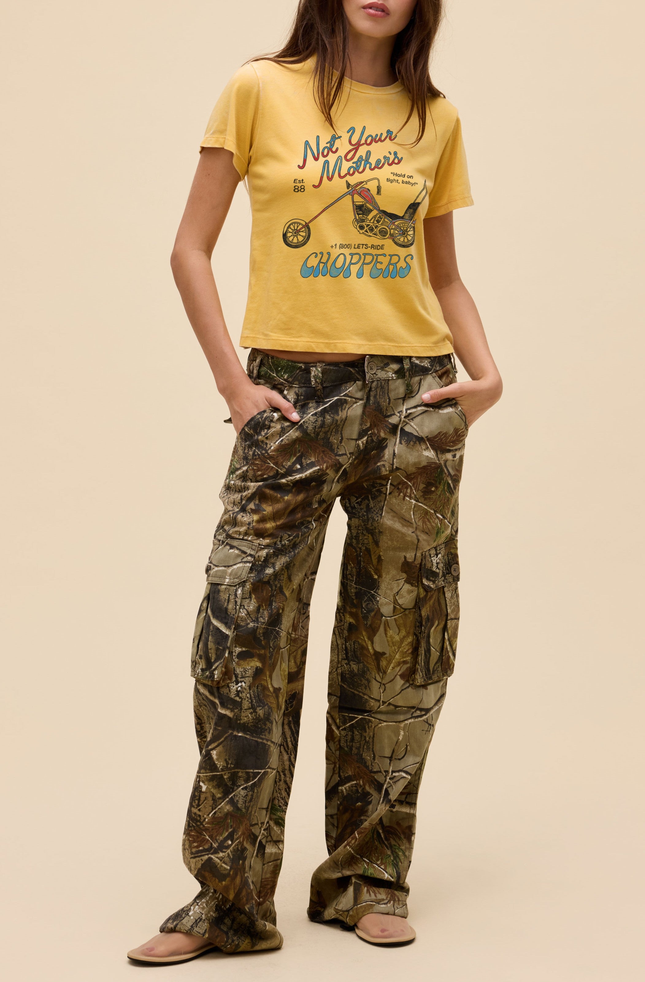 A person stands against a beige background, wearing Daydreamer LA's Not Your Mothers Choppers yellow Burnout Vintage Tee in a shrunken fit and loose camo cargo pants, hands in pockets, and brown sandals on their feet.