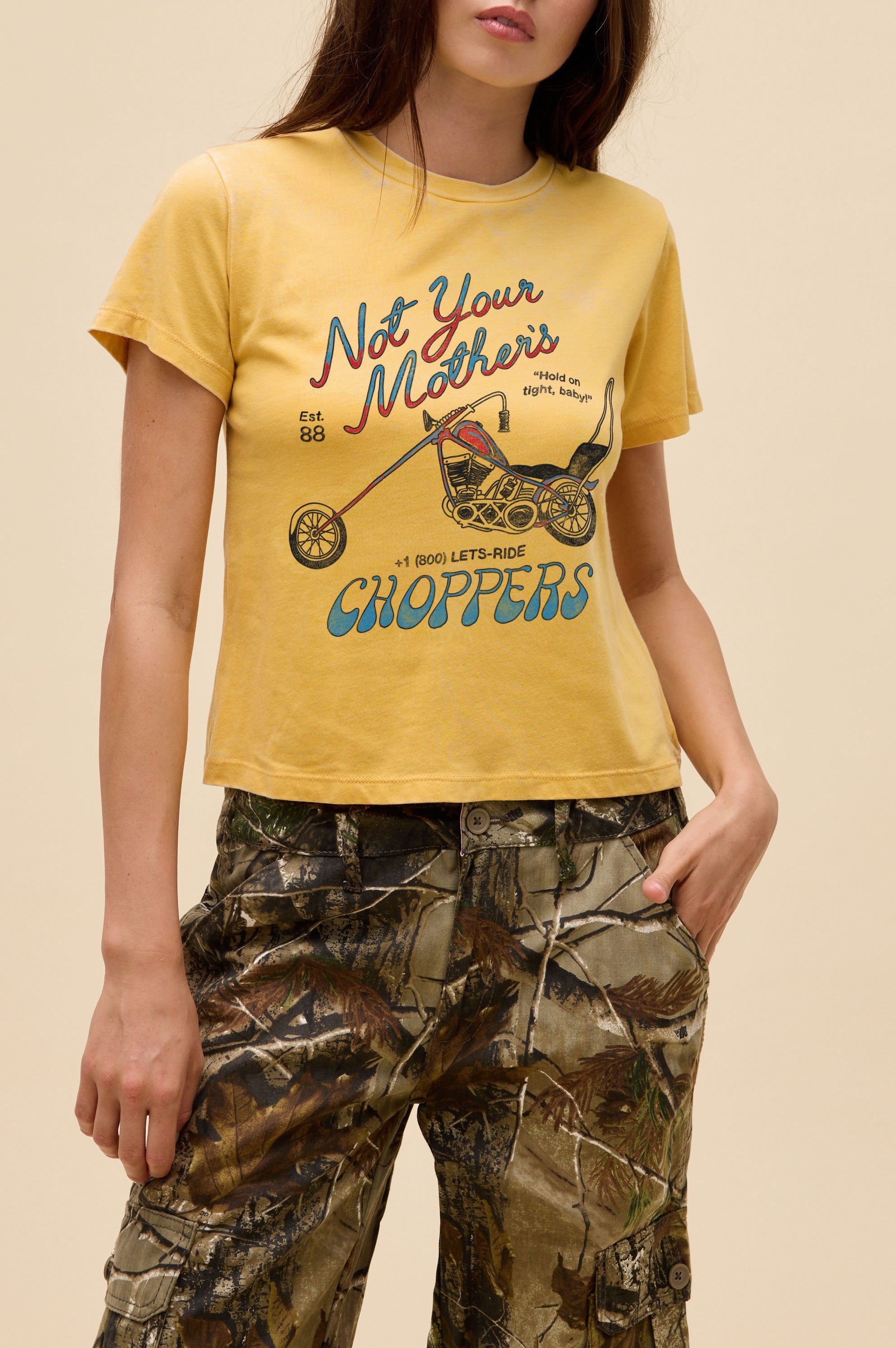 A woman stands with her hands in her pockets, wearing the yellow Daydreamer LA "Not Your Mothers Choppers" tee paired with camouflage cargo pants, set against a plain background.