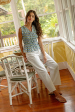 A woman with long brown hair sits on a white chair in a sunlit room, wearing the THML Blue Printed Smocked Top with white pants and sandals. The room features yellow walls, large windows, and nearby tables and chairs.