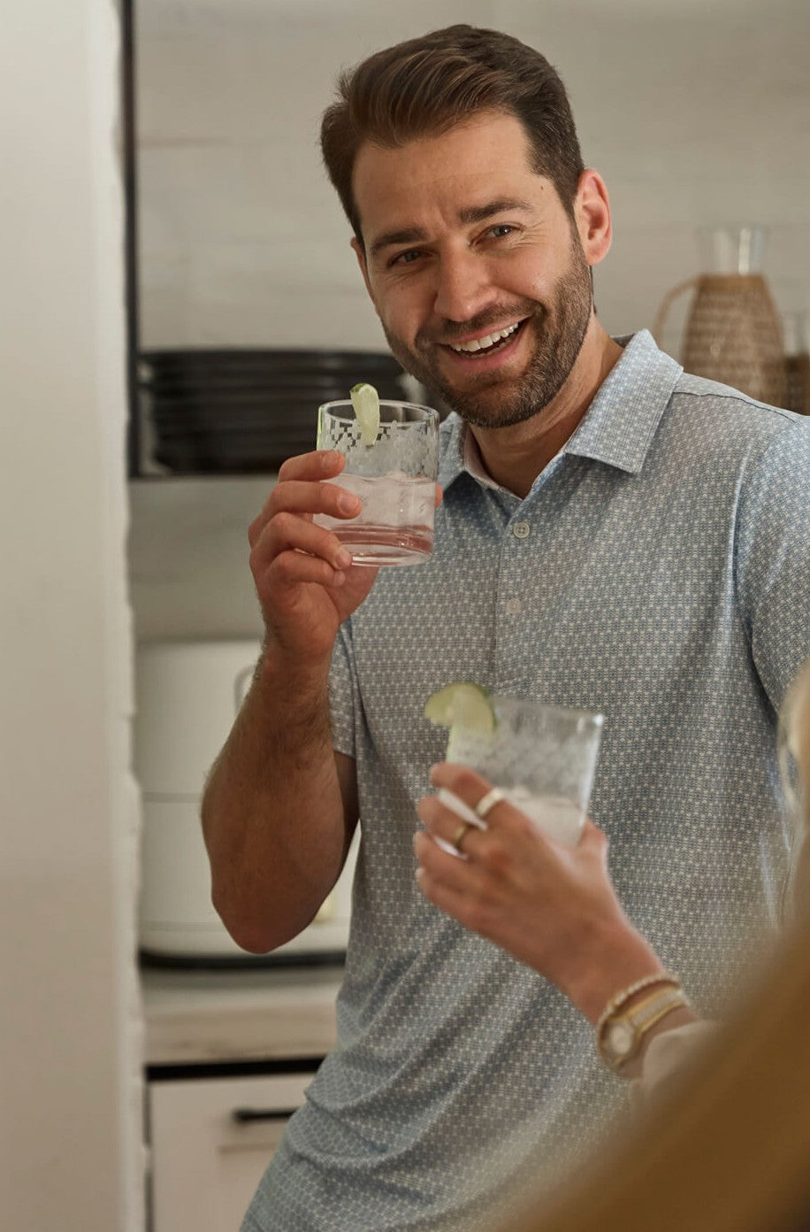 A man in the Toes on the Nose Clubhouse Polo Tequila Therapy in light blue smiles and holds a lime-garnished drink, standing in a kitchen next to another person with a similar beverage.