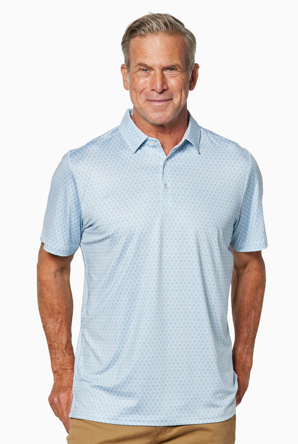 A middle-aged man smiles in a Toes on the Nose Clubhouse Polo Tequila Therapy, a light blue quick-dry short-sleeve shirt with a subtle pattern, paired with tan pants against a white background.