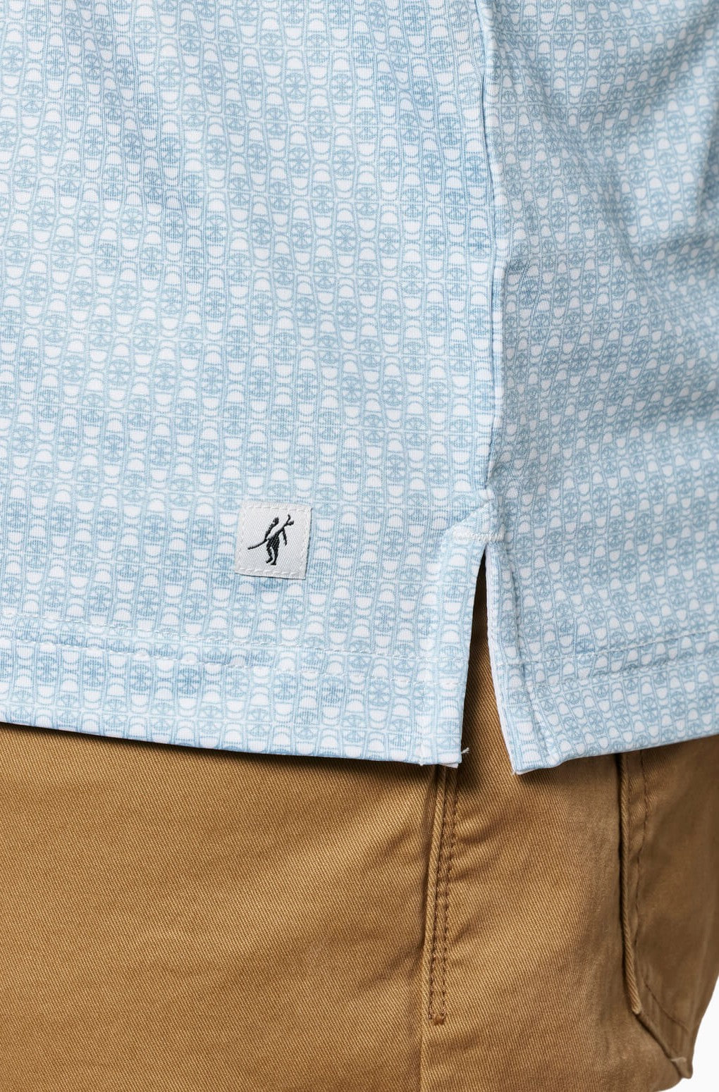 A close-up of a person wearing light brown pants and the Toes on the Nose Clubhouse Polo Tequila Therapy, a light blue patterned shirt with a black figure logo tag near the hem, designed for quick-dry performance and comfort.
