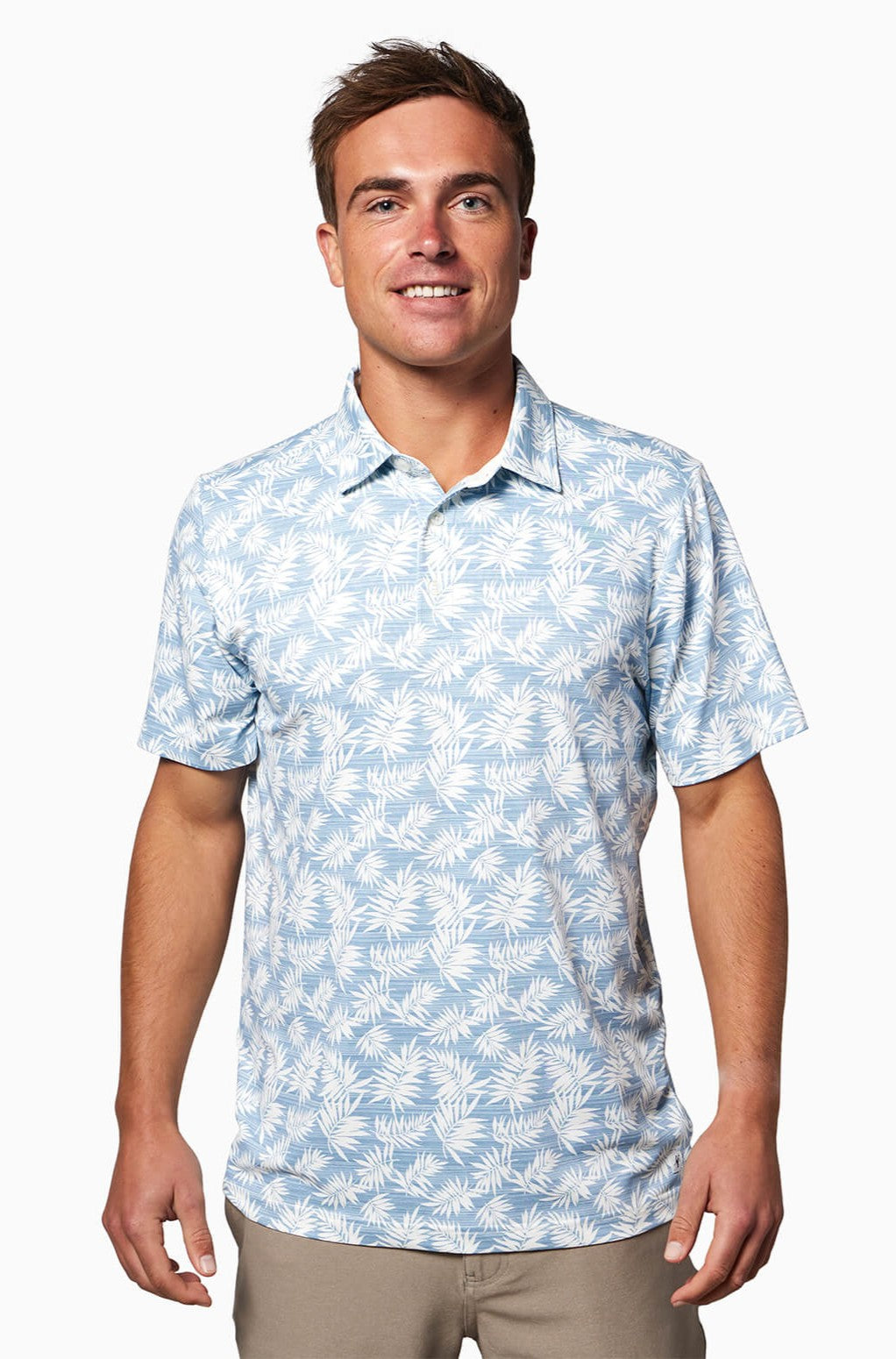 A man with short brown hair smiles in the Toes on the Nose Clubhouse S/s Polo, a light blue short-sleeve shirt featuring a white palm leaf pattern and comfortable moisture-wicking poly stretch fabric. He stands against a plain white background.
