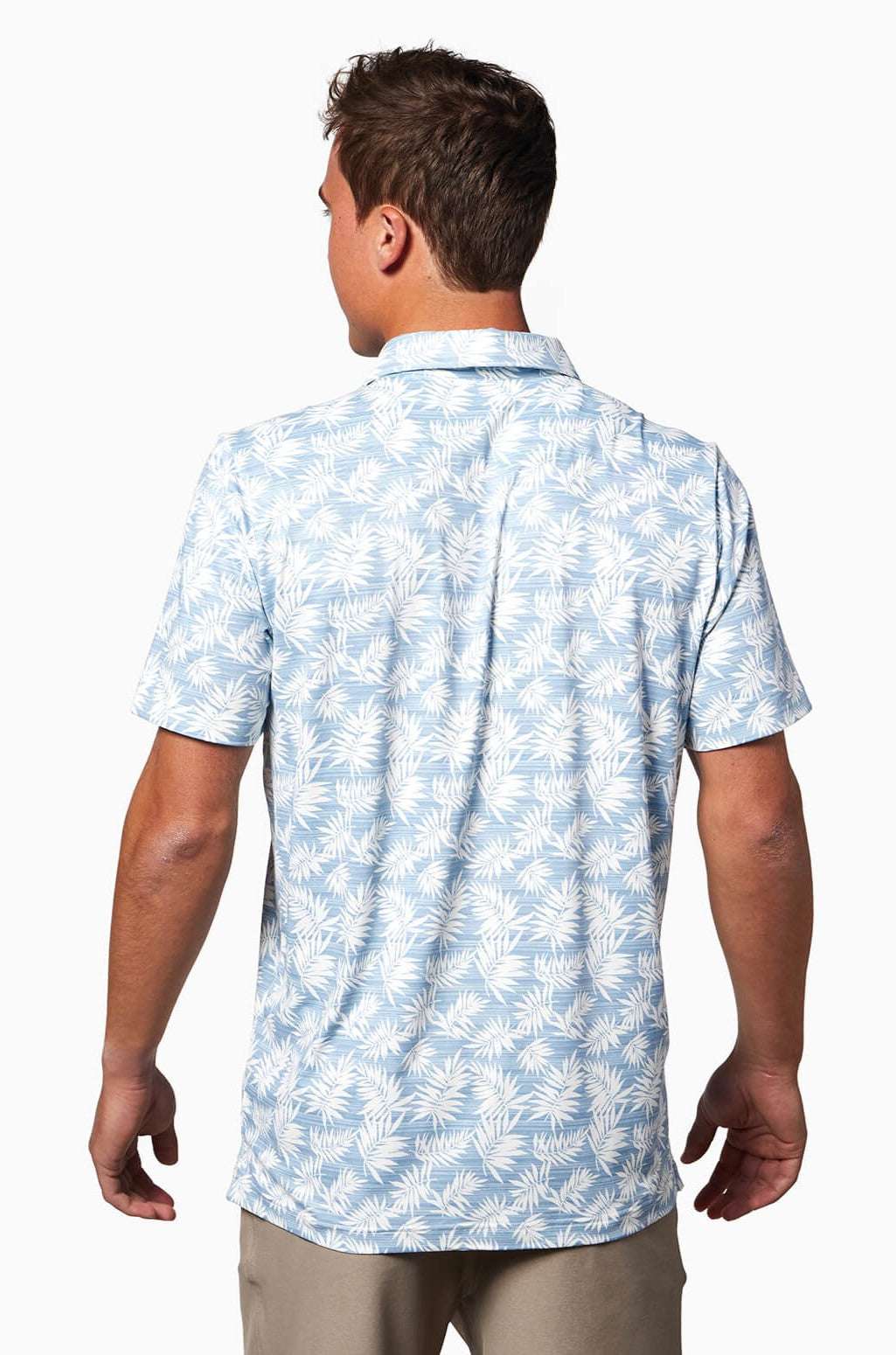 A man is shown from behind wearing the Toes on the Nose Clubhouse S/s Polo, a light blue, quick-dry short-sleeve shirt with a white palm leaf pattern, paired with beige pants against a plain white background.
