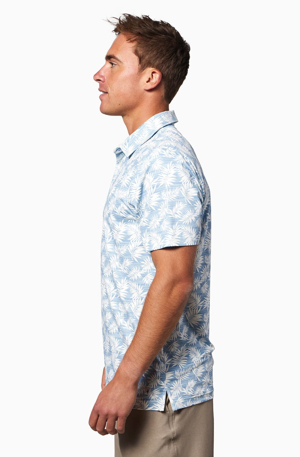 A man in profile wears the Toes on the Nose Clubhouse S/s Polo—a light blue, quick-dry, short-sleeve shirt with a white palm leaf pattern—and beige pants, standing against a plain white background.