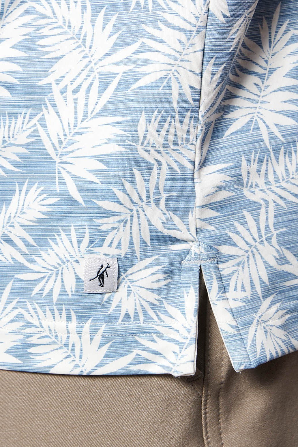 Close-up of the Clubhouse S/s Polo by Toes on the Nose—a light blue, moisture-wicking, quick-dry shirt with a white tropical leaf pattern and a logo tag near the hem—paired with beige pants.