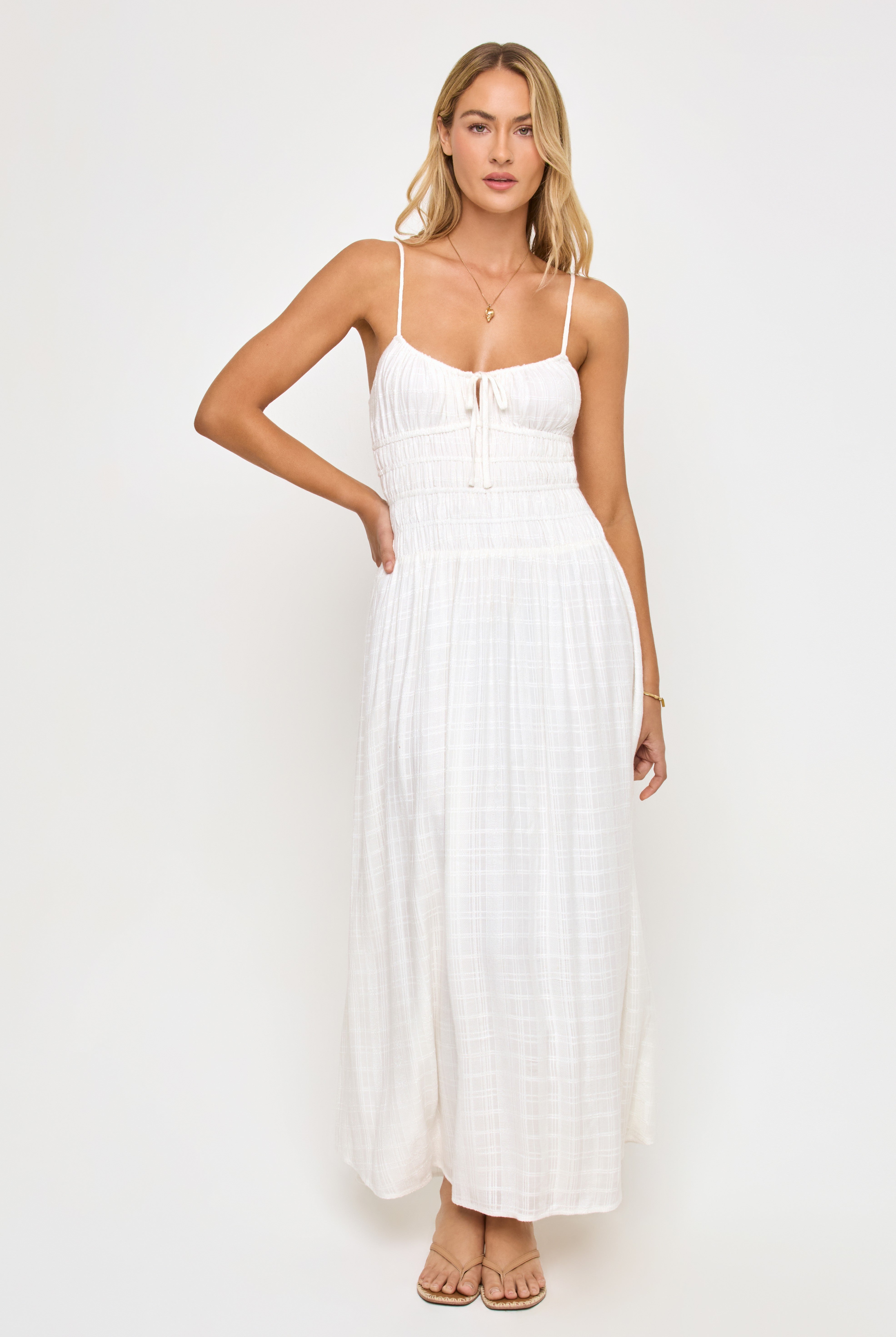 A woman with long blond hair models the LSpace Tala Maxi Dress, a sleeveless white spaghetti strap maxi with a fitted ruched bodice. She stands against a plain white background, wearing sandals and minimal jewelry.