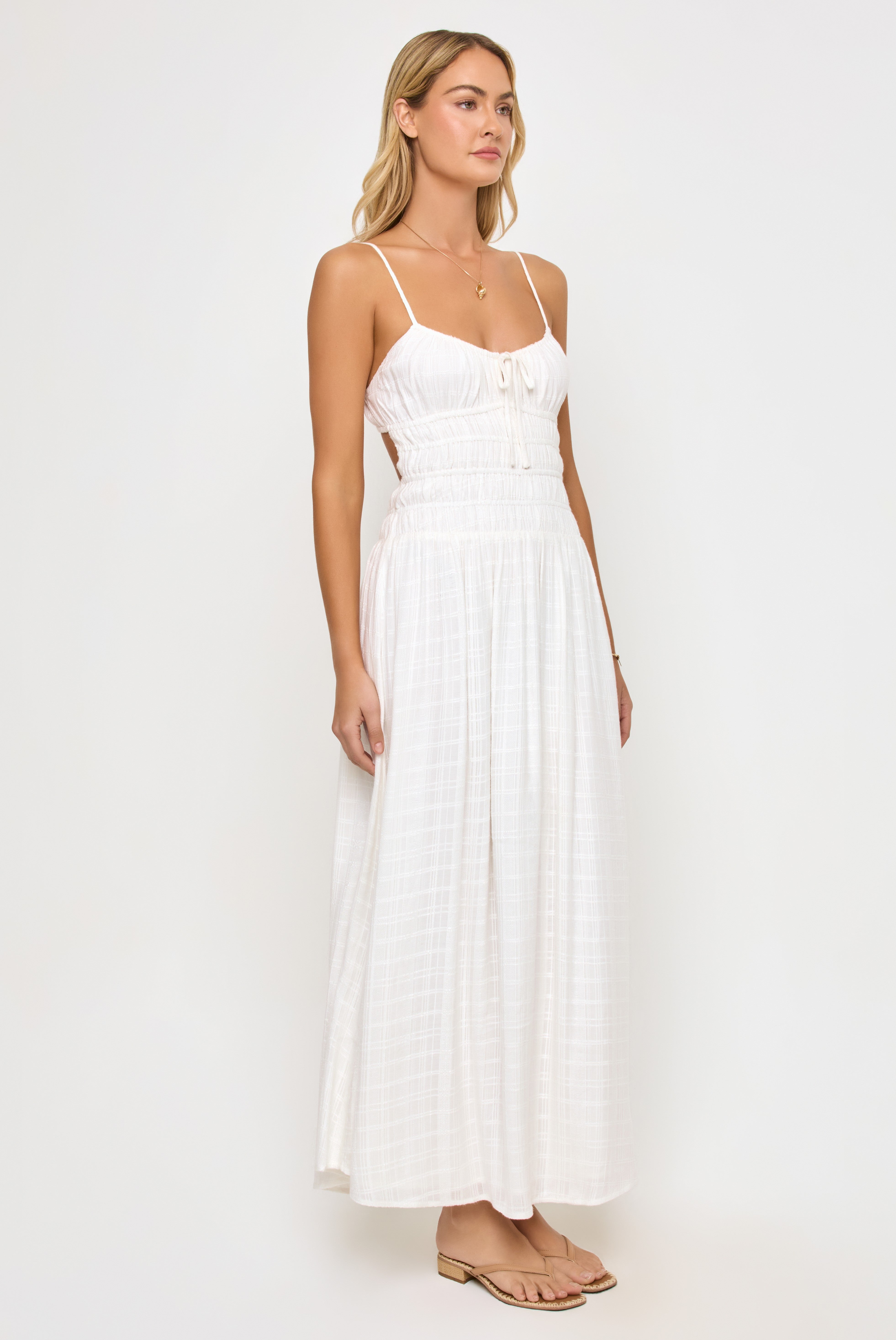A woman with light hair stands against a white background wearing the LSpace Tala Maxi Dress—a sleeveless, white spaghetti strap maxi dress with a smocked waist—paired with flat sandals and a simple necklace.