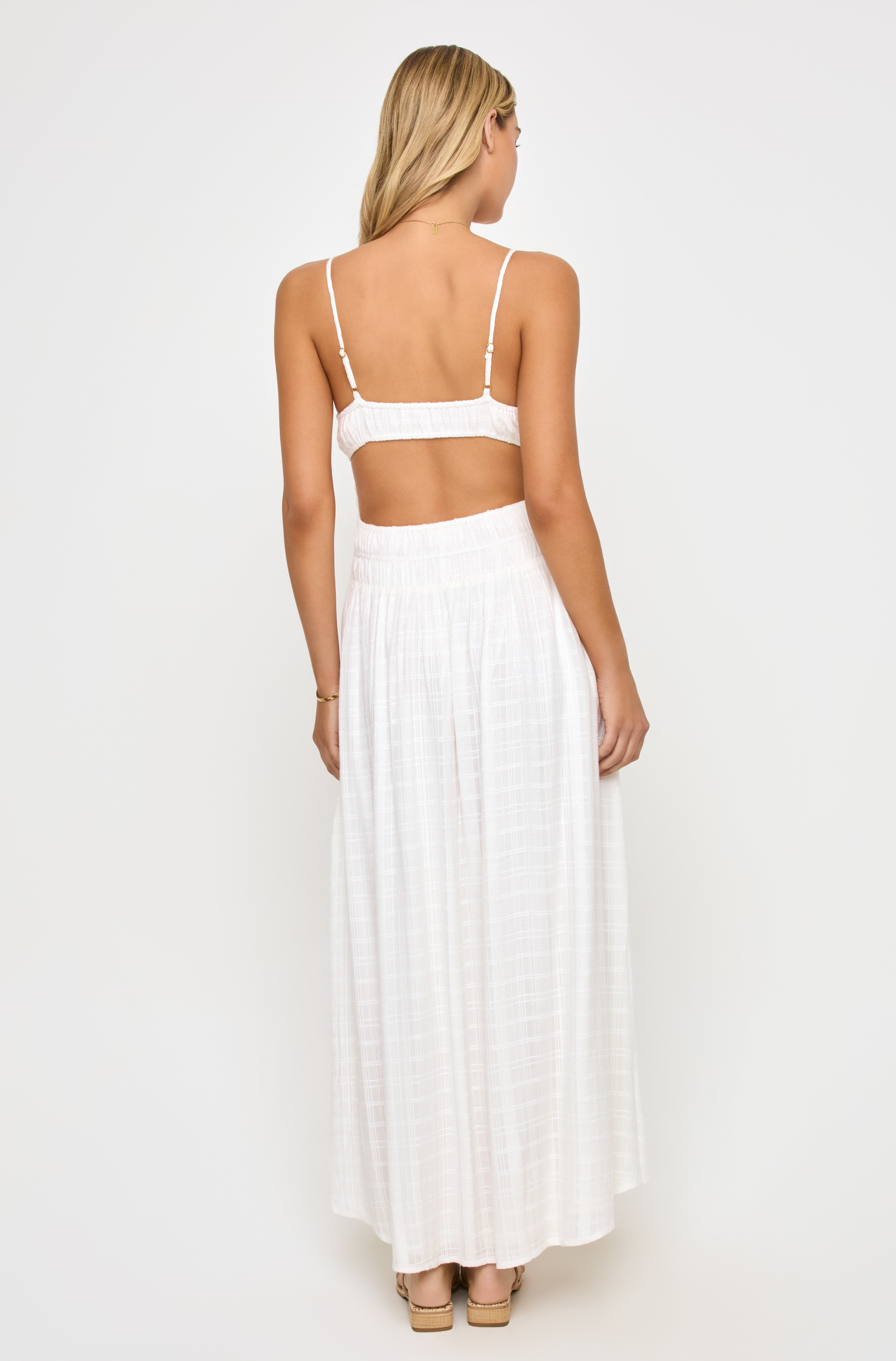 A woman with blonde hair, facing away from the camera against a plain background, wears the LSpace Tala Maxi Dress—a white spaghetti strap dress featuring a smocked back and cutout detail.