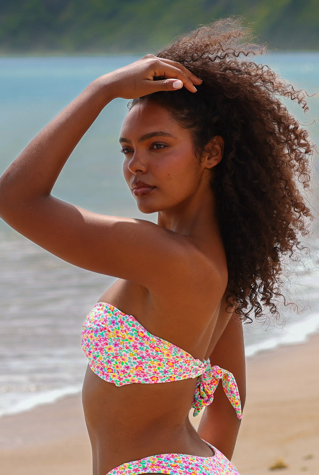 A woman in a Kulani Kinis Field Trip Strapless Bandeau Bikini Top stands on a sandy beach, shielding her eyes with one hand as the ocean and a green hillside stretch out behind her.