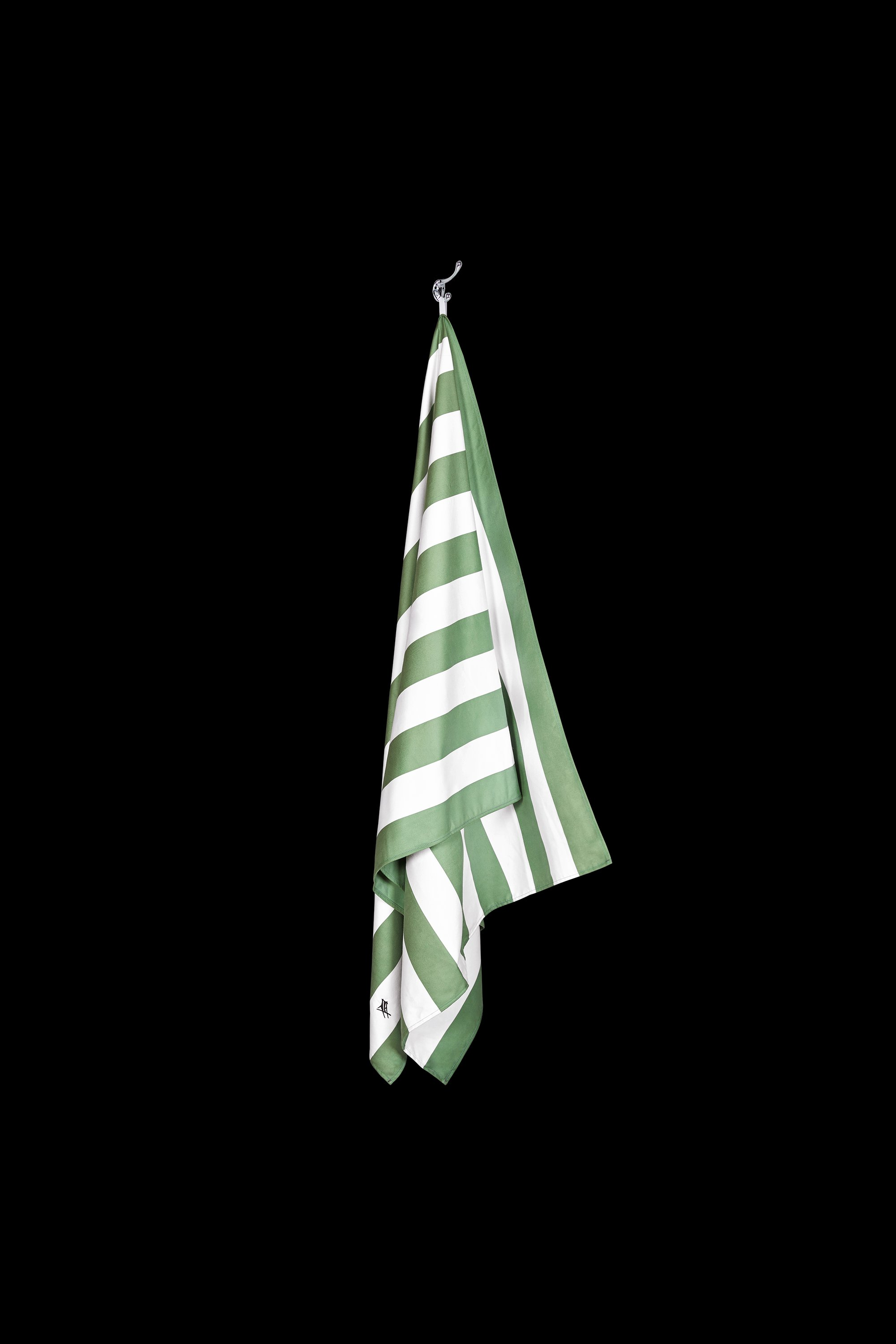 The Cayman Olive Stripe Towel by South Moon Under, featuring green and white stripes, hangs from a hook against a solid black background.