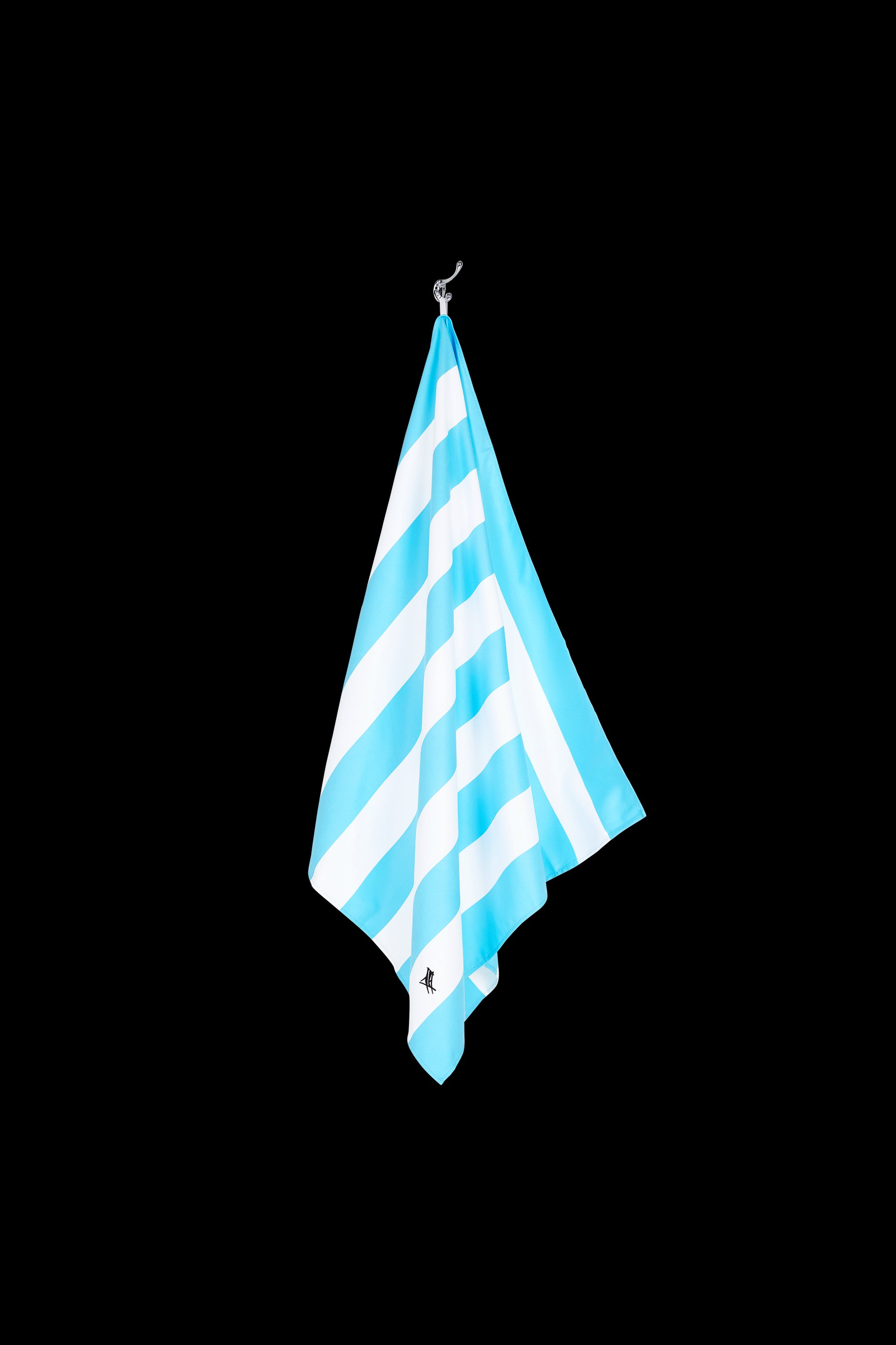 The South Moon Under Tulum Blue Large Stripe Towel, made with quick-dry microfibre, features blue and white diagonal stripes and a small logo at the bottom, shown hanging from a hook against a black background.