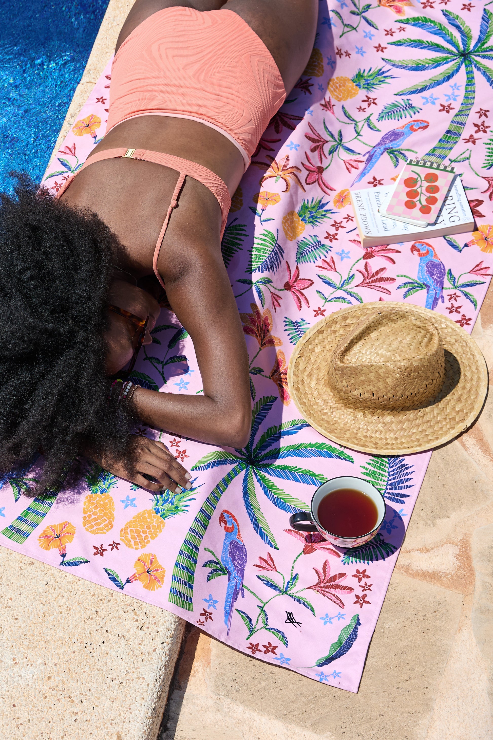A person in a peach swimsuit relaxes poolside on the Pink Paradise Large Towel by South Moon Under, a lightweight, quick-dry microfibre towel with palm trees, parrots, and flowers; nearby are a straw hat, cup of tea, and two books.