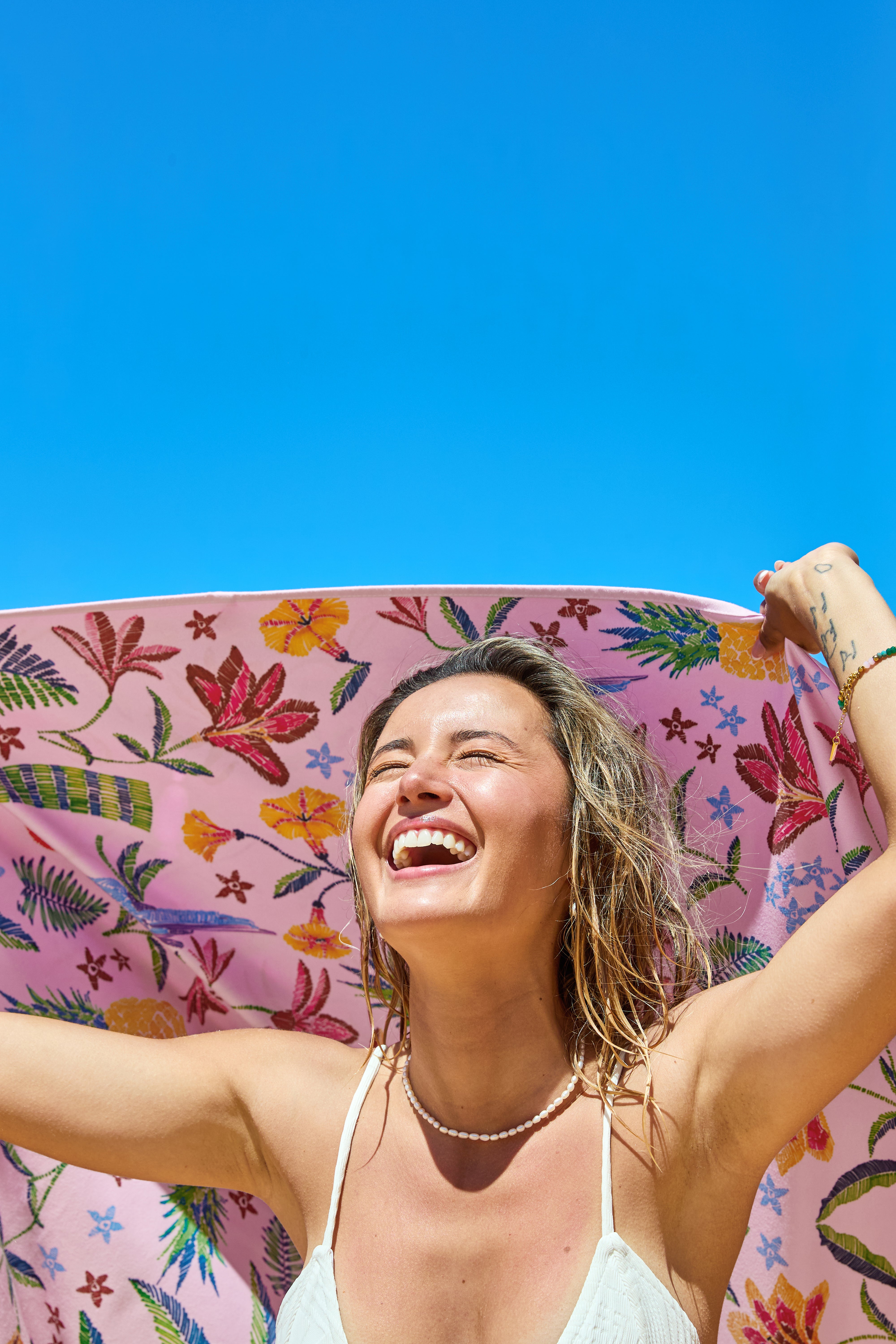 A smiling woman holds the South Moon Under Pink Paradise Large Towel in Pink Multi above her head under a blue sky, wearing a white top and pearl necklace, enjoying the sunny day.